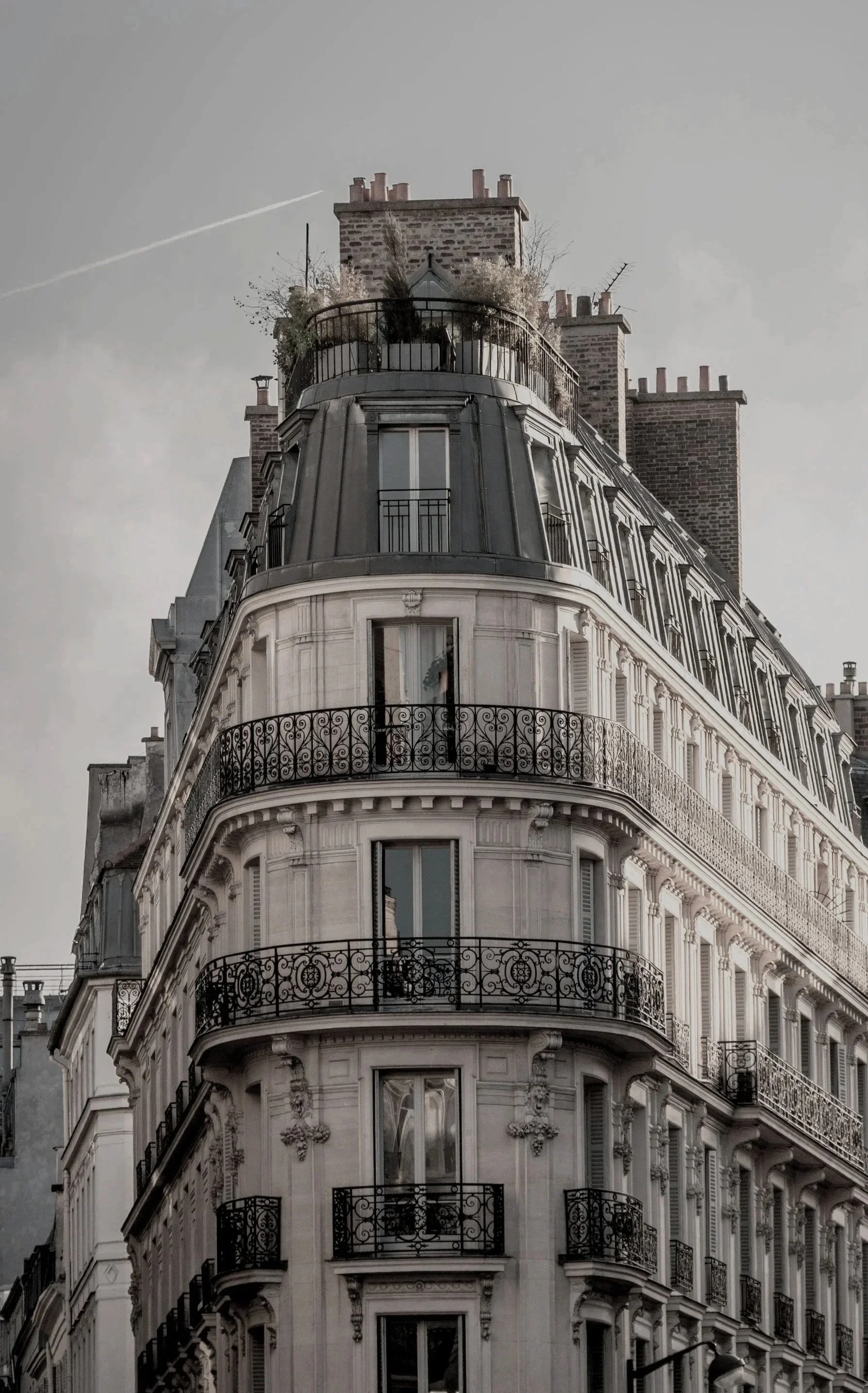 A classic Parisian building with ornate white stone facade, multiple balconies with wrought iron railings, and tall windows. The top features a mansard roof with dormer windows and a rooftop terrace with greenery.