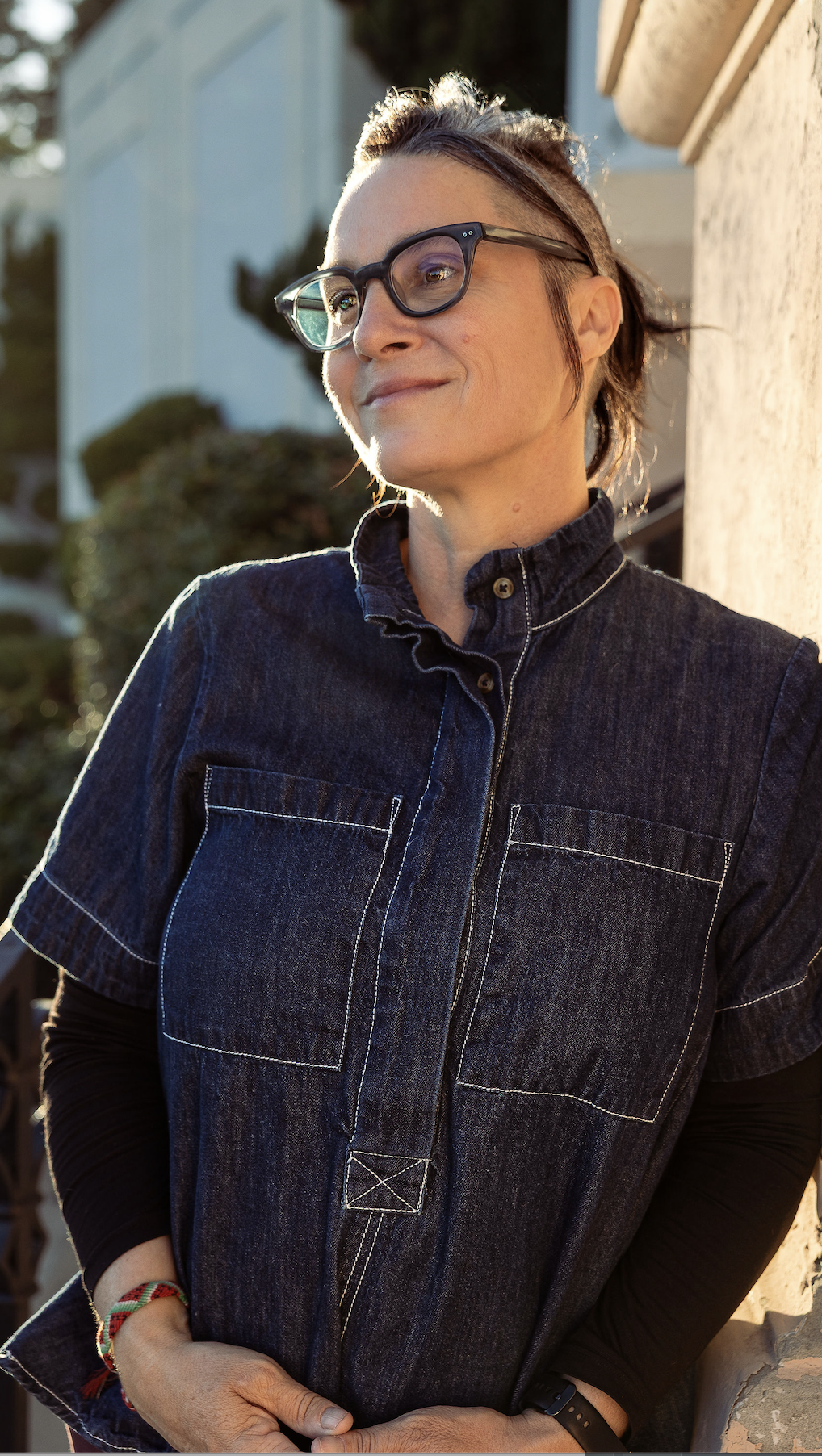 A woman with short brown hair, wearing glasses and a dark denim jacket, standing outdoors near a wall in warm sunlight.