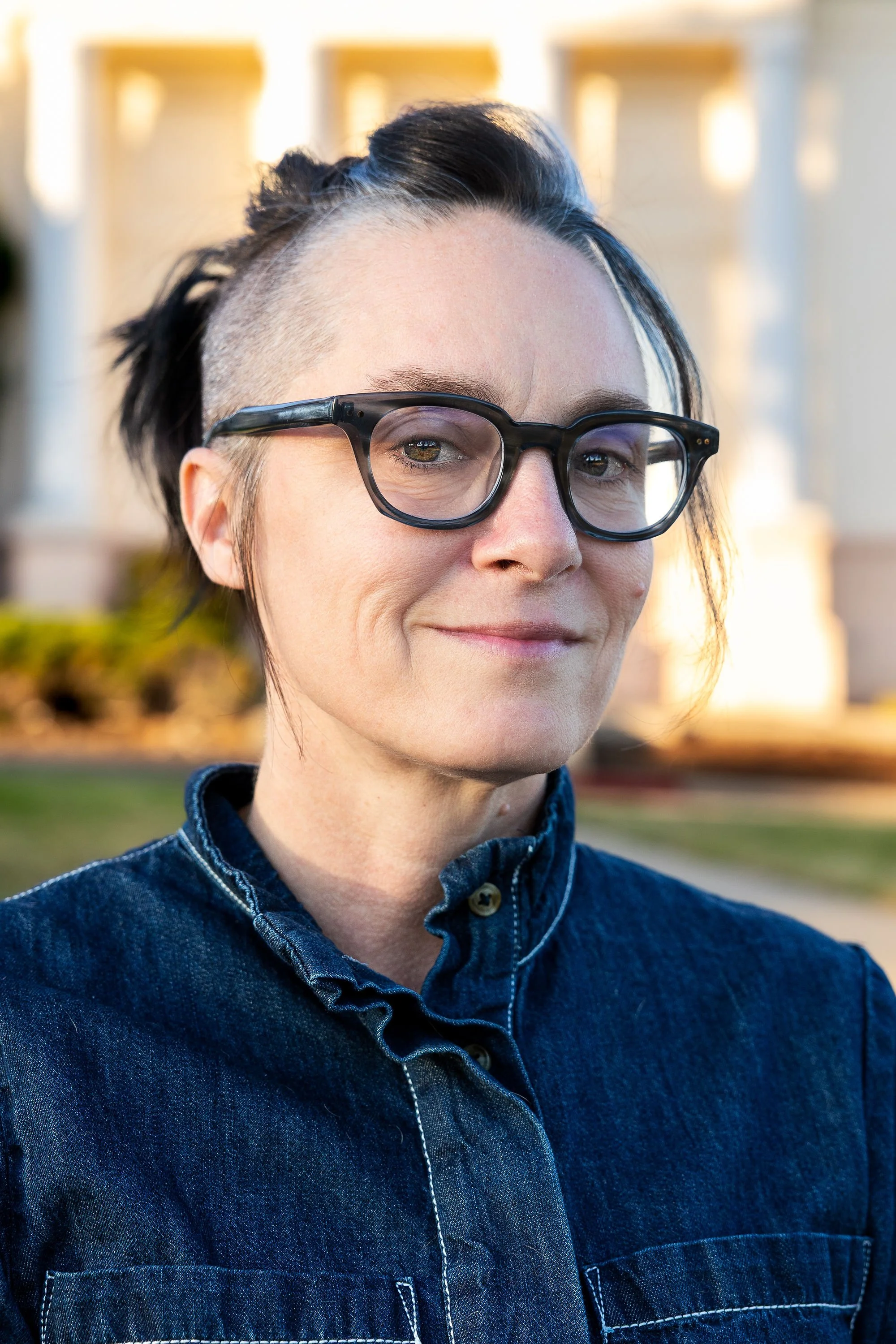 Close-up of a person with black and gray hair, wearing black-rimmed glasses and a denim shirt, standing outdoors with a blurred background of light-colored buildings.