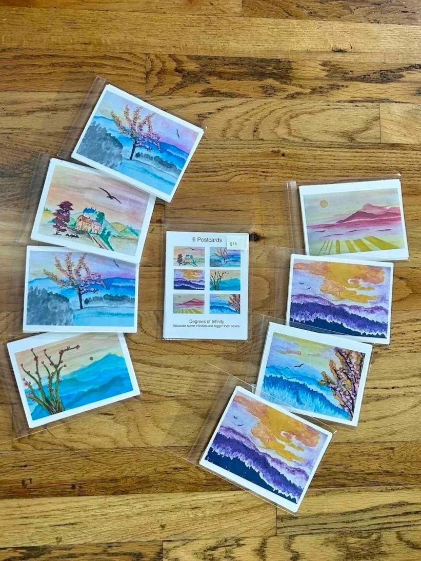 Six postcards featuring watercolor landscape paintings with mountains, trees, lakes, and sunsets, displayed on a wooden surface. A small flyer in the center advertises 6 postcards for $15.