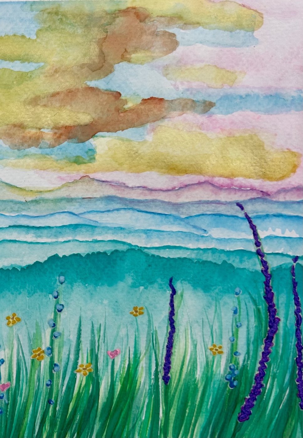 Watercolor painting of a landscape with a sky filled with colorful clouds, layered mountains, and a green field with yellow and purple flowers and tall grasses.