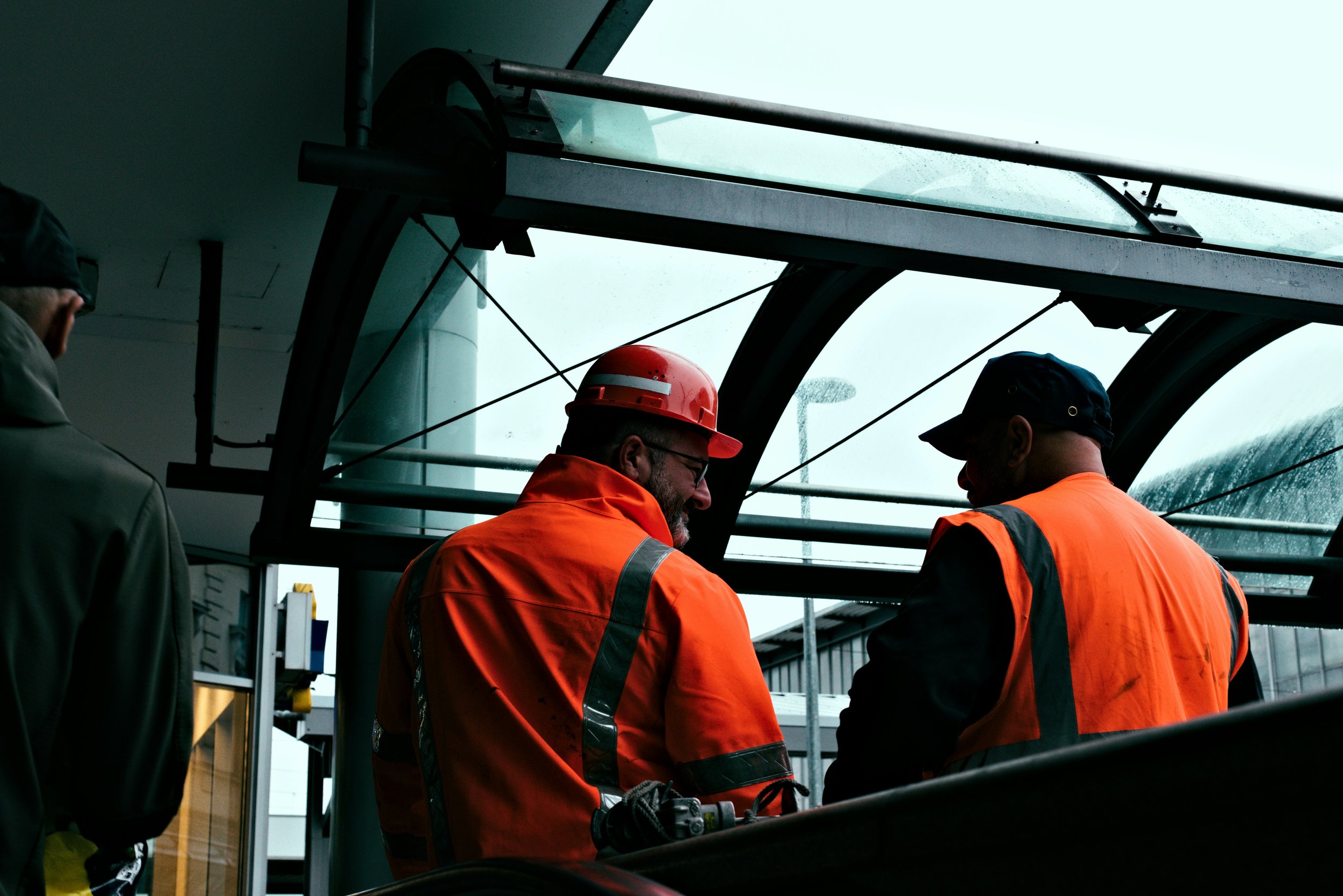Construction workers in orange safety vests and helmets standing on scaffolding at an airport or transportation hub, with glass and metal structures around them.