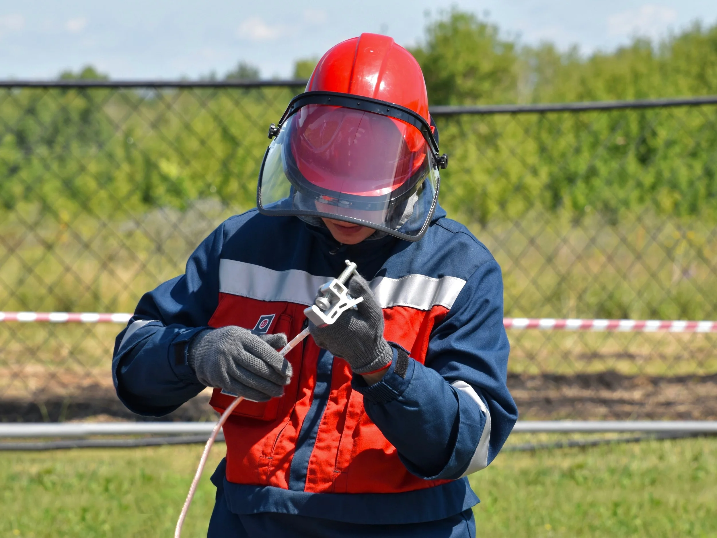 Person wearing a red helmet and face shield working outdoors with a chain-link fence and green trees in the background.