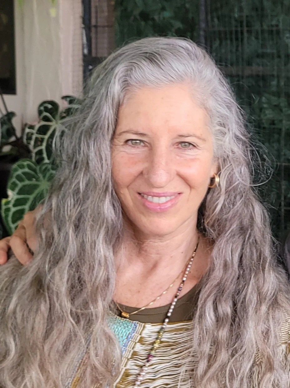 Close-up of a woman with long gray curly hair, smiling, wearing gold earrings and necklaces, with plants in the background.