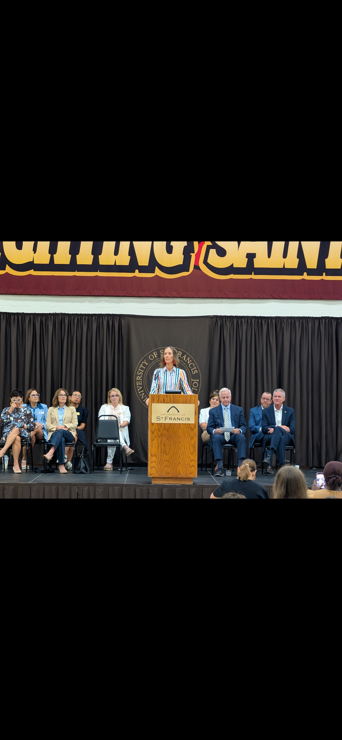 A woman giving a speech at a podium during an event at the University of San Francisco, with eight seated individuals behind her on a stage, including men and women in professional attire, under a banner that reads 'Learning & Social'.