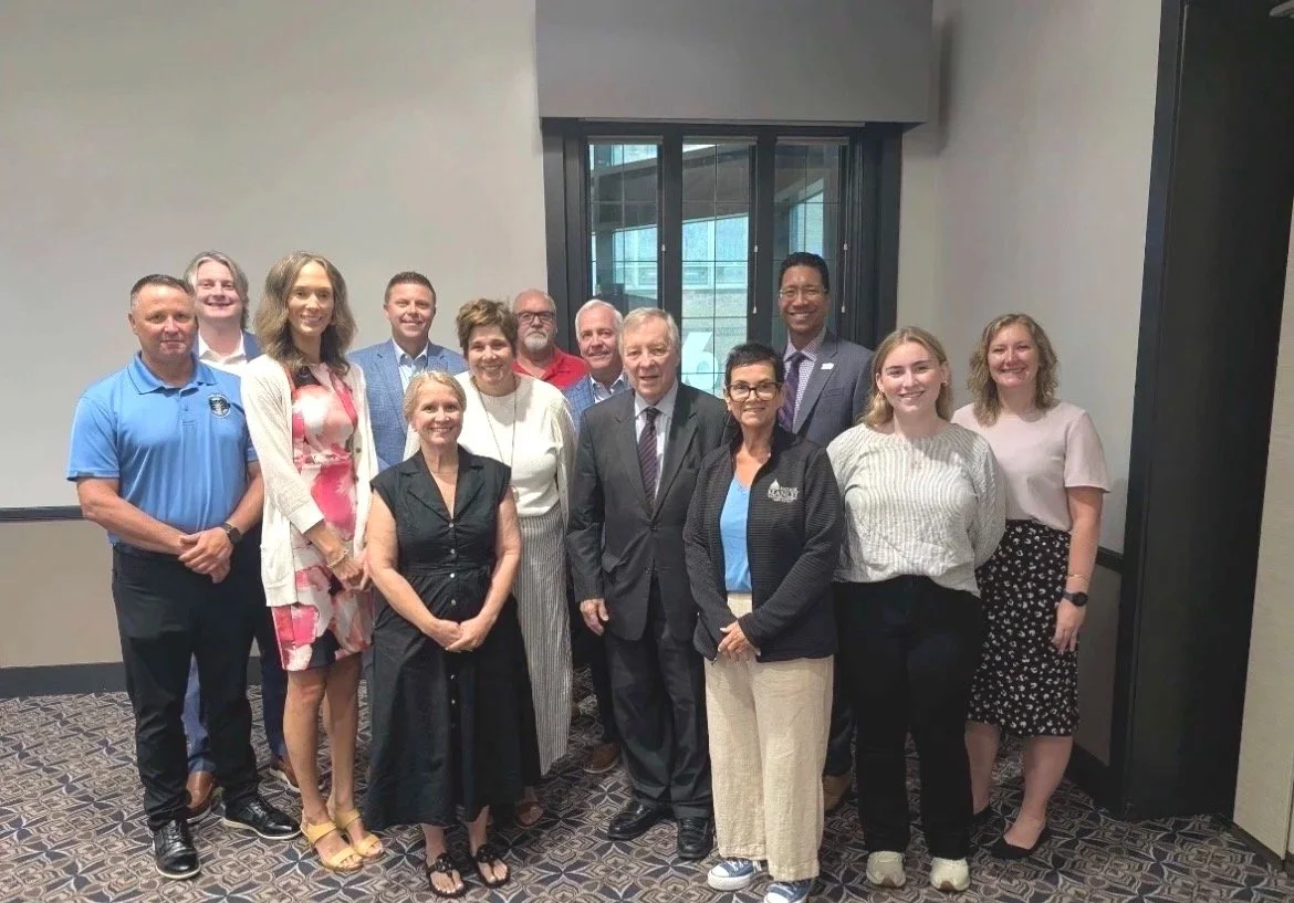 A group of fifteen people standing together inside a conference room, posing for a photo, with some smiling and others looking at the camera.