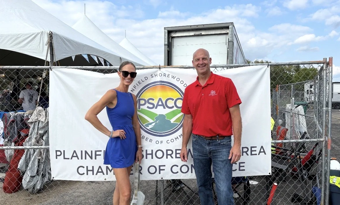 A woman in a blue dress and sunglasses standing next to a man in a red polo shirt, both smiling, at an outdoor event with a sign that says "Plainfield Shorewood Area Chamber of Commerce" and the PSACO logo in the background.