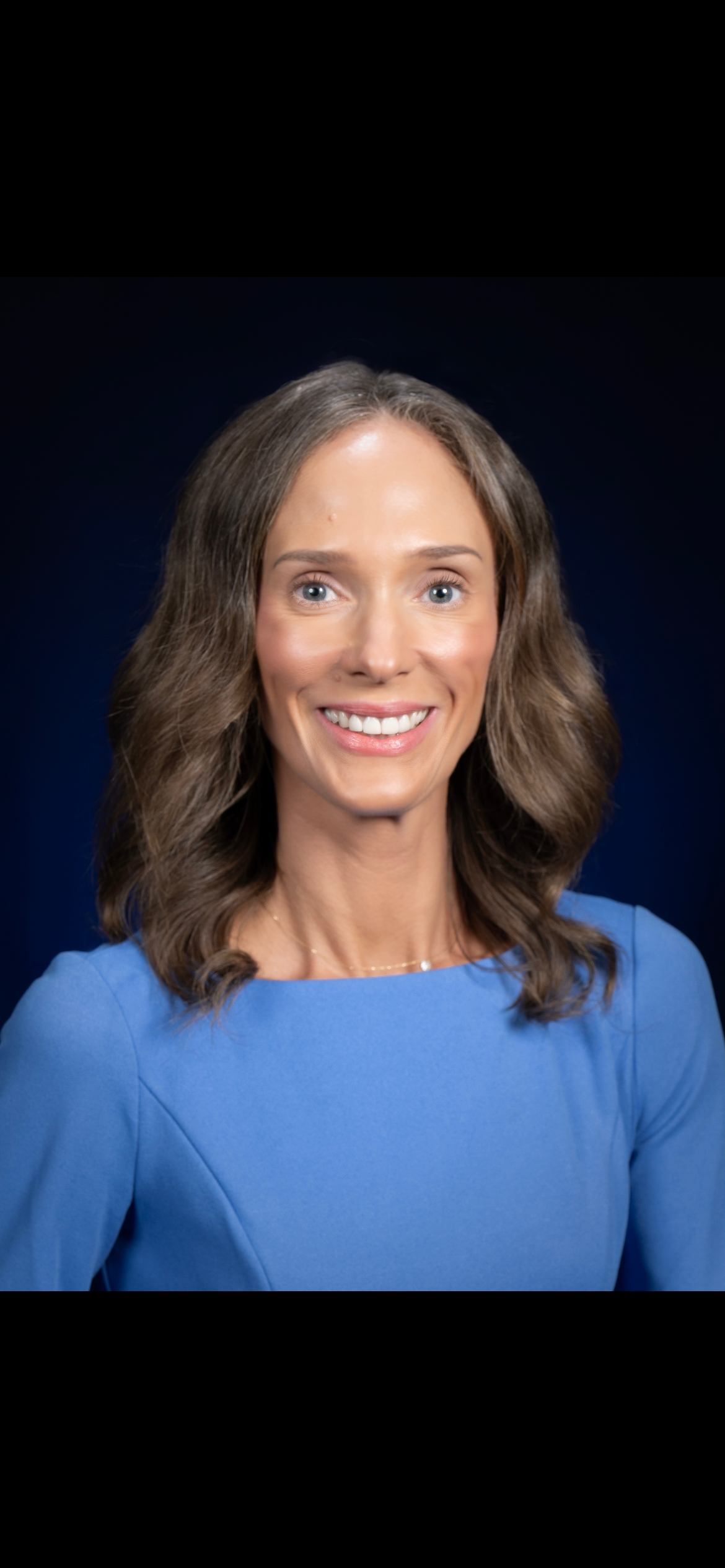 A smiling woman with shoulder-length wavy brown hair, wearing a blue dress, against a dark blue background.