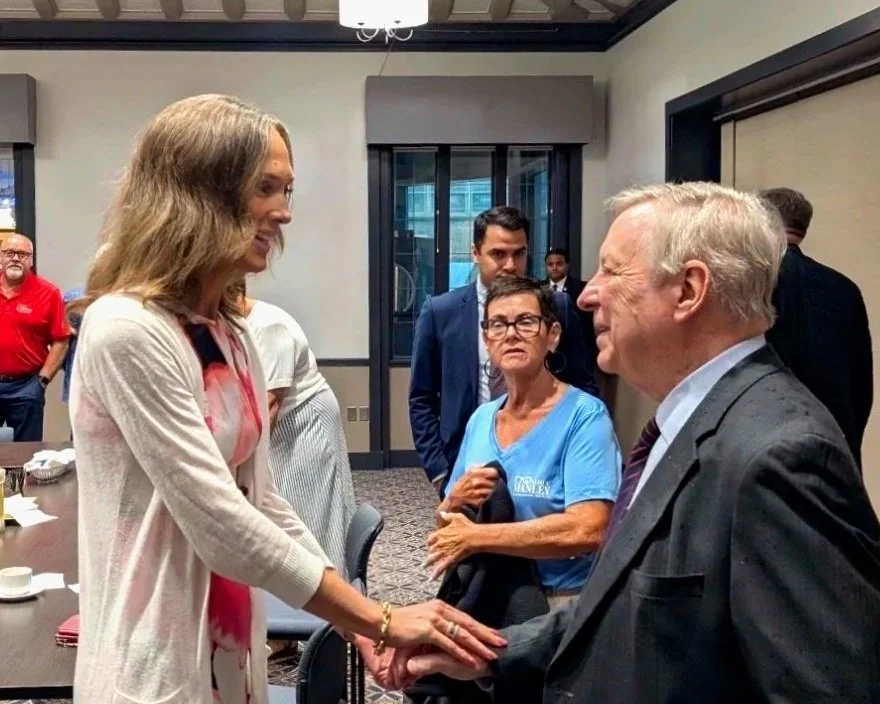 A woman with shoulder-length light brown hair interacts with an elderly man in a suit, greeting him with a handshake and a gentle touch on his arm, inside a conference room with several people observing.