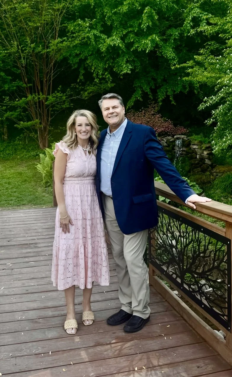 Keith and Rachel Leitner standing on a wooden deck outdoors surrounded by green trees, with a small water feature behind them.
