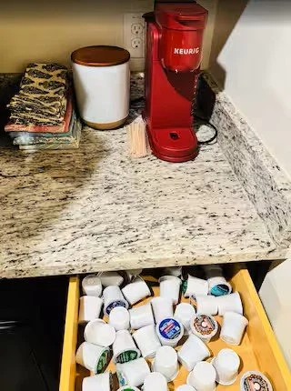 A kitchen counter with a red Keurig coffee maker, a container with a lid, a stack of napkins, and a drawer filled with assorted coffee pods.