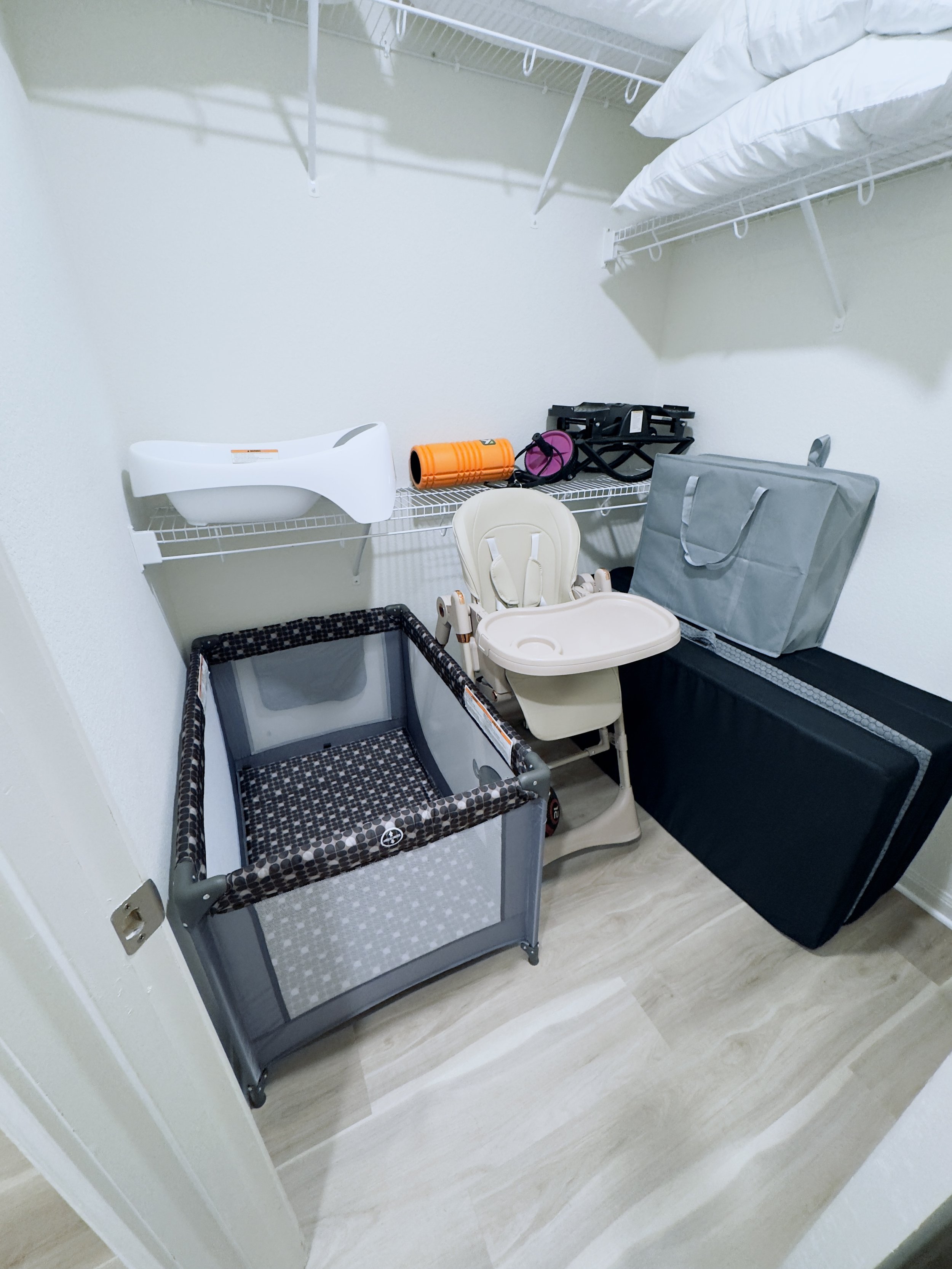 A walk-in closet with white wire shelves on the walls, containing pillows and clothes. On the floor, there is a baby crib with a leopard print fabric, a white high chair, and a gray storage bag. The closet has light-colored flooring.