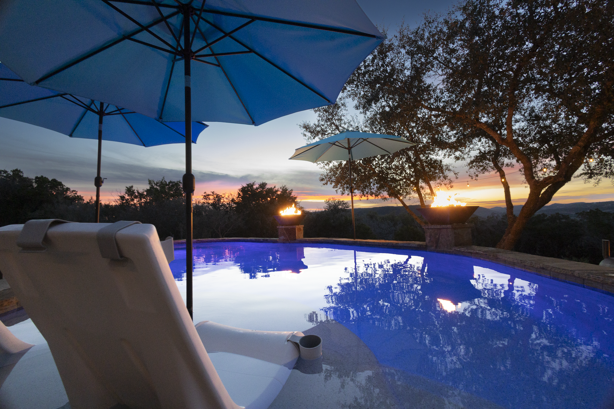 A backyard pool area at sunset with umbrellas, lounge chairs, and a fire feature, surrounded by trees and scenic mountains in the distance.
