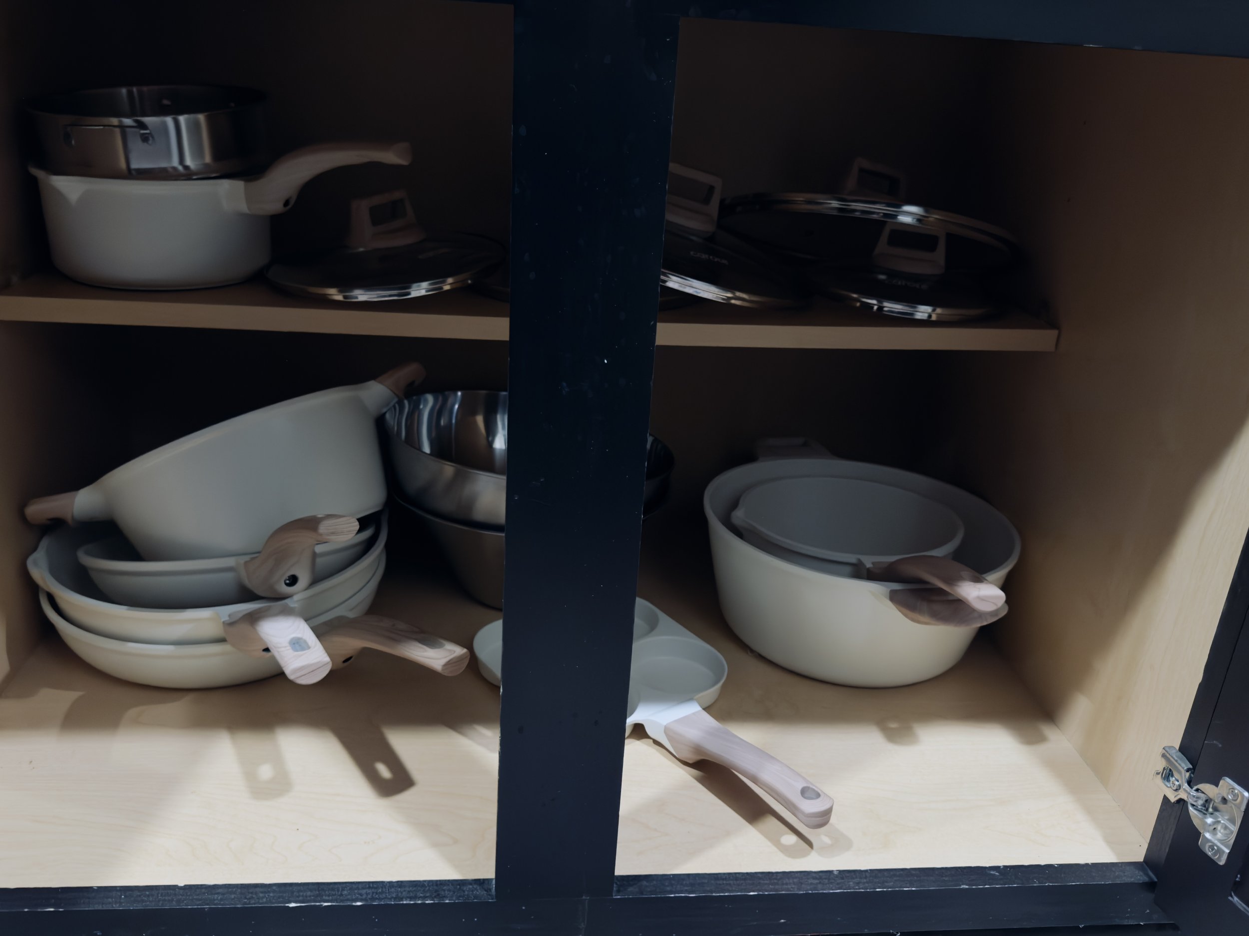 Open kitchen cabinet with a white ceramic teapot with a wooden handle, metal pots and various bowls on shelves inside.