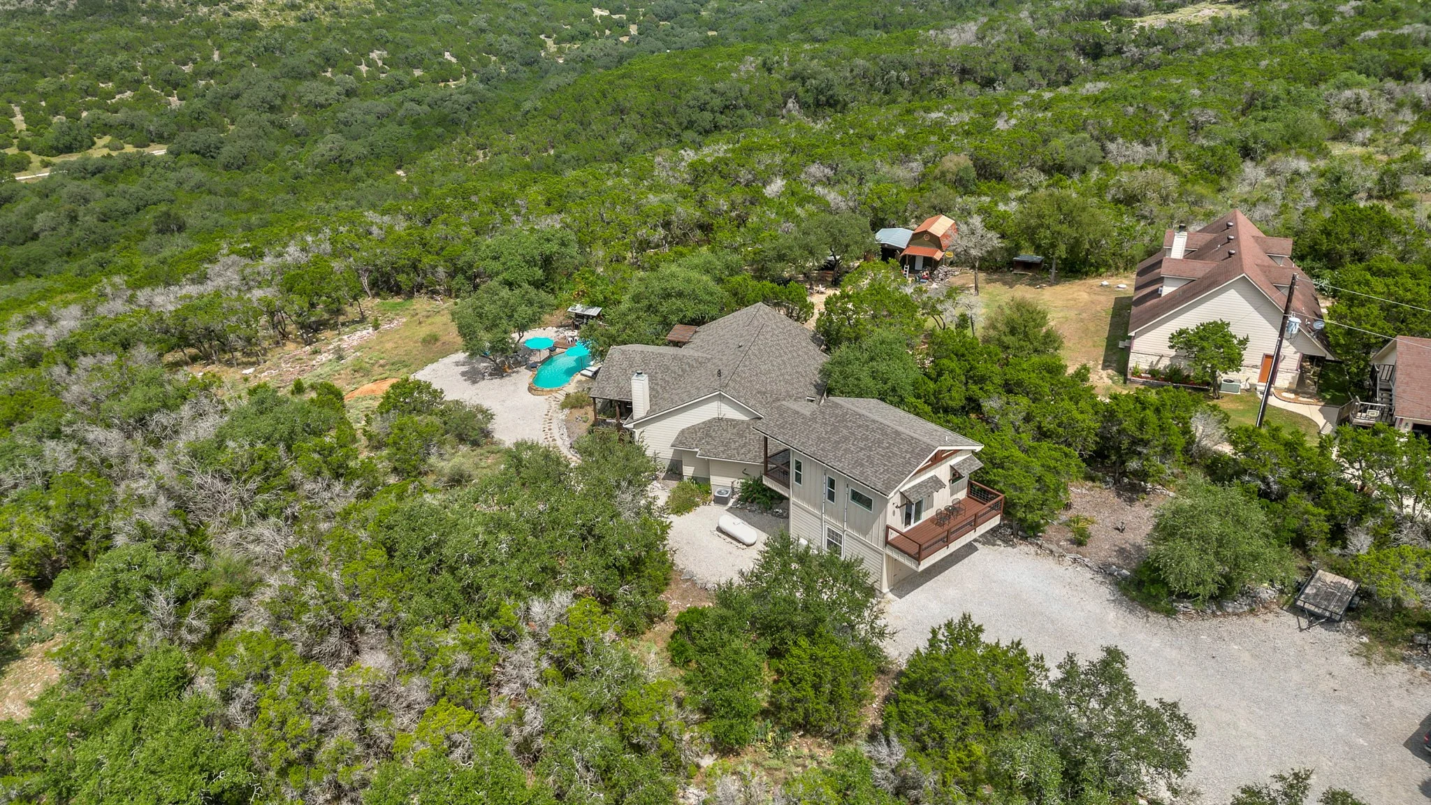 Aerial view of a residential area surrounded by trees, featuring houses with varied rooftops, a swimming pool, and outdoor living spaces.