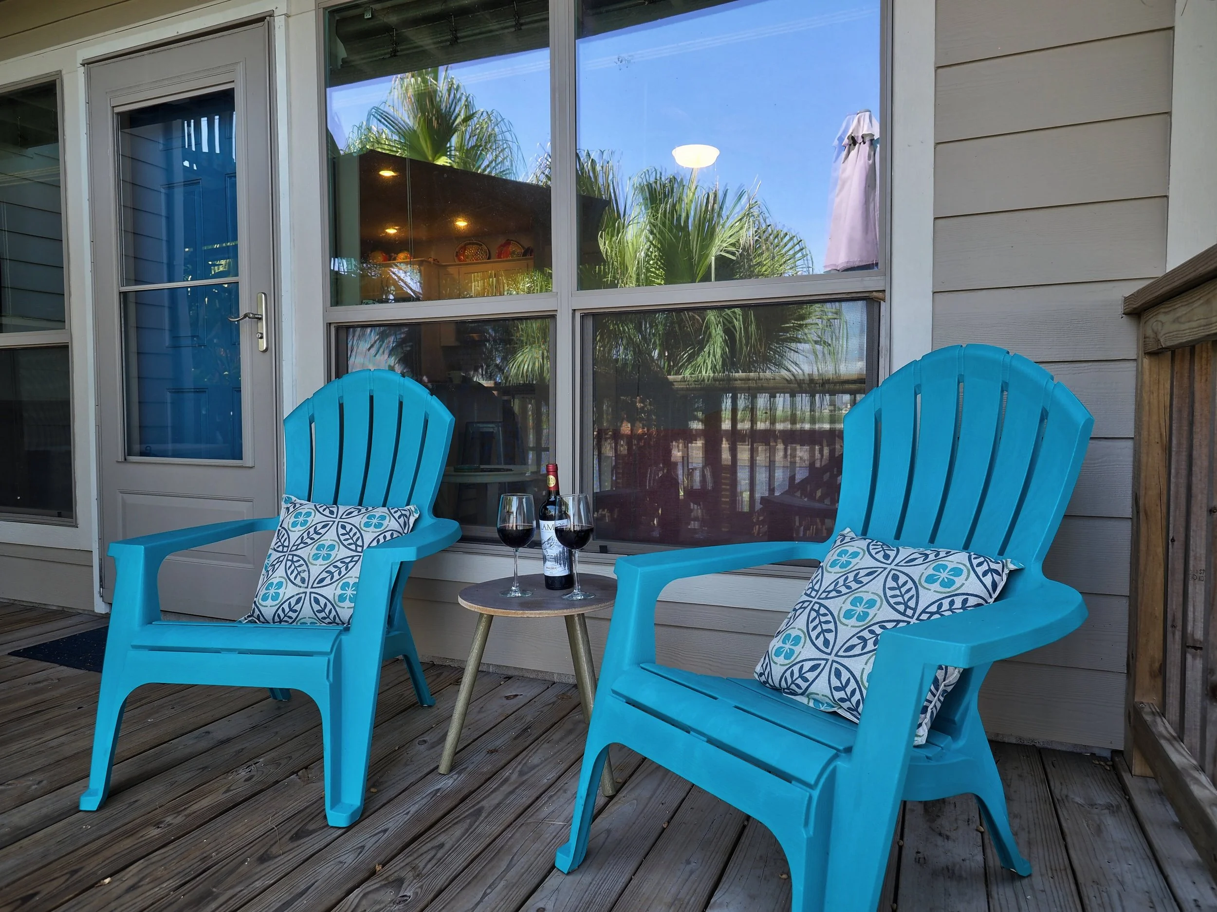 Two bright blue patio chairs with patterned cushions and a small round table holding two glasses of red wine and a bottle, on a wooden deck outside a house with large front window and a door.