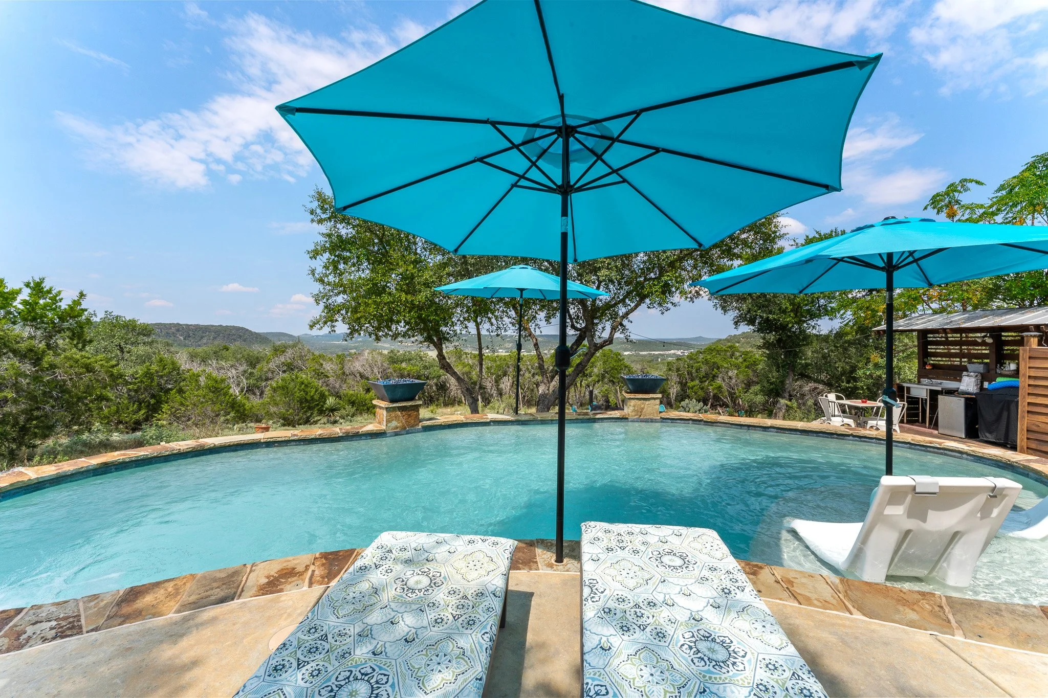 Swimming pool with two lounge chairs, a large blue patio umbrella, and a smaller blue umbrella in a backyard with trees and a scenic view of hills under a partly cloudy sky.