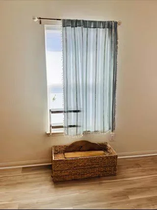 A window with sheer blue curtains in a room with light-colored walls and wooden flooring, and a wicker storage bench underneath the window.
