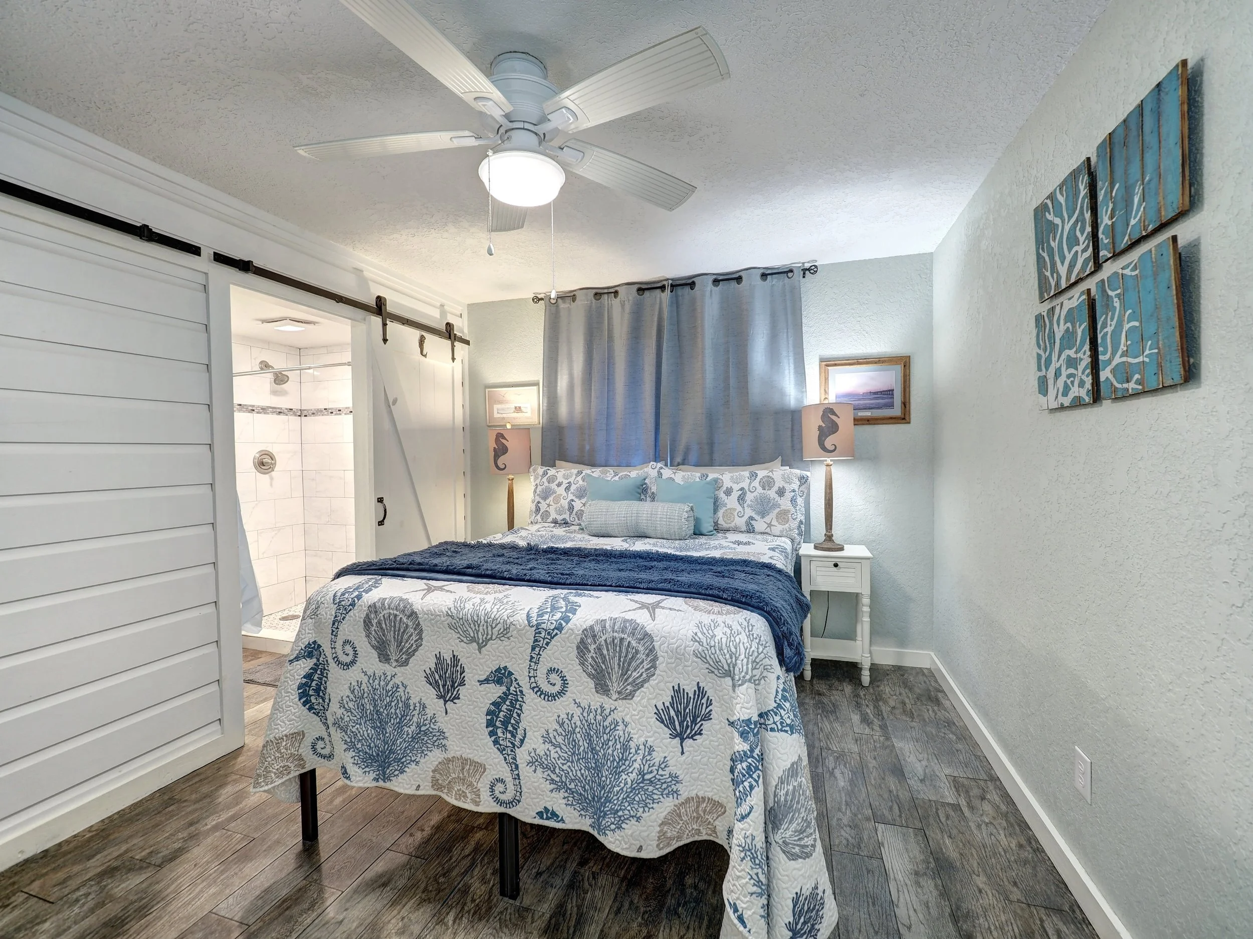 A bedroom with a white ceiling fan, a bed with blue and white sea life themed bedding, and nautical decor. An open door shows a bathroom with a shower.