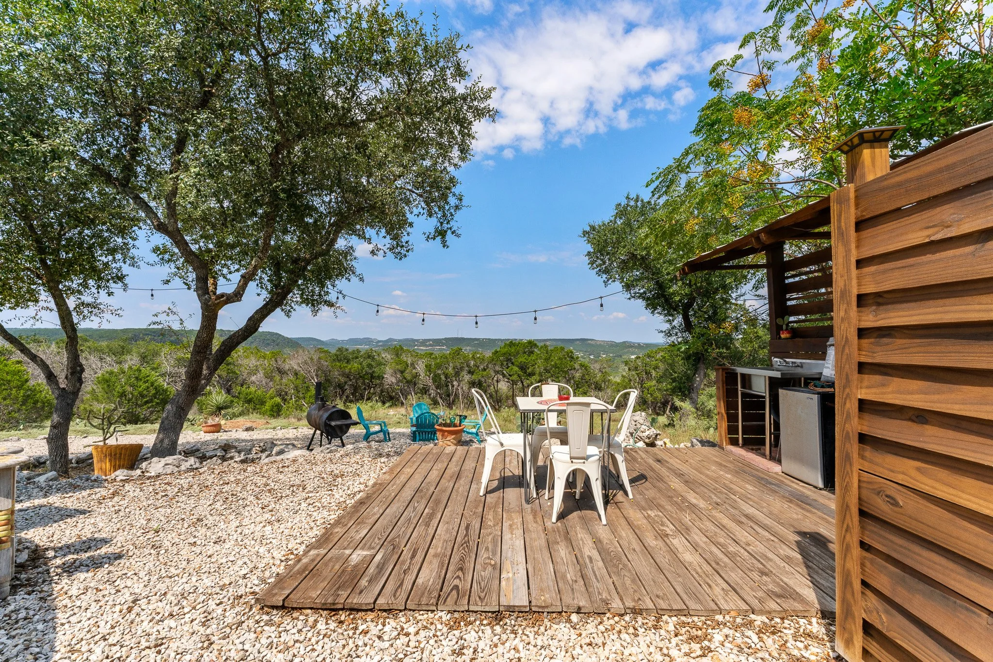 An outdoor patio with a wooden deck, white and turquoise chairs, a round table, a grill, and string lights, surrounded by trees under a blue sky with clouds.