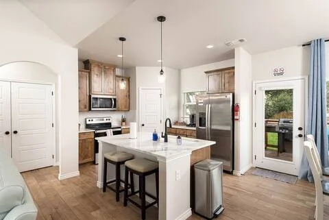 Kitchen with wooden cabinets, stainless steel refrigerator, white island with seating, pendant lights, and a door leading outside.