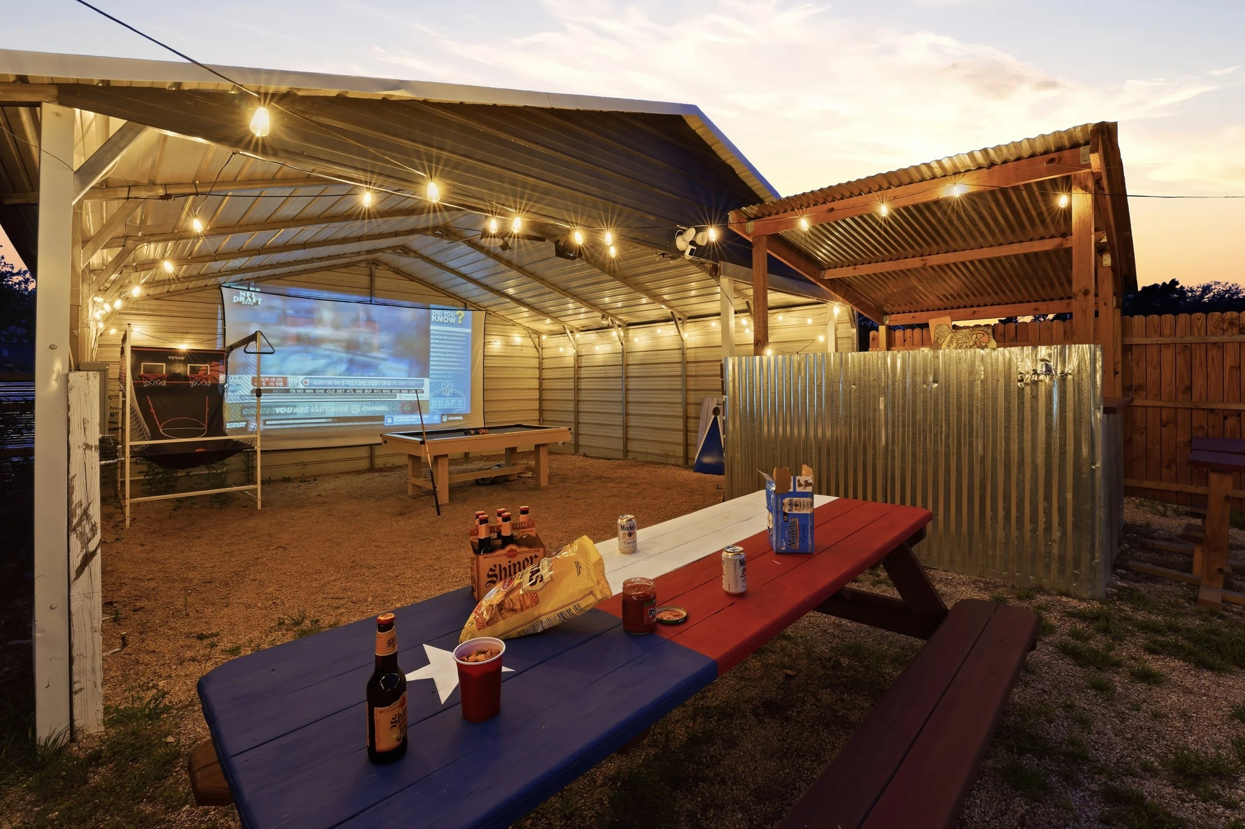 An outdoor covered patio area at sunset with string lights. There is a picnic table with snacks and beverages, and a large outdoor screen showing a sports broadcast. In the background, there is a wooden privacy fence and a small wooden structure.