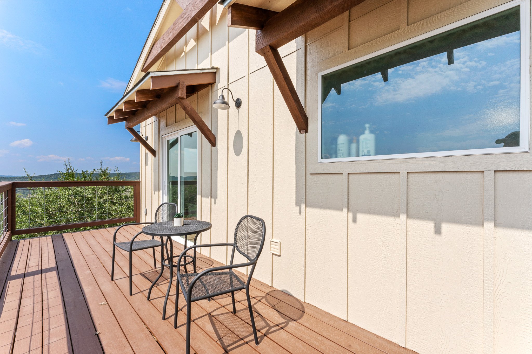 Photo of an outdoor balcony with a wooden deck, black chairs around a small round table, and a railing with a view of trees and hills in the background. The building exterior has beige siding, large windows, and decorative wooden awnings with metal s