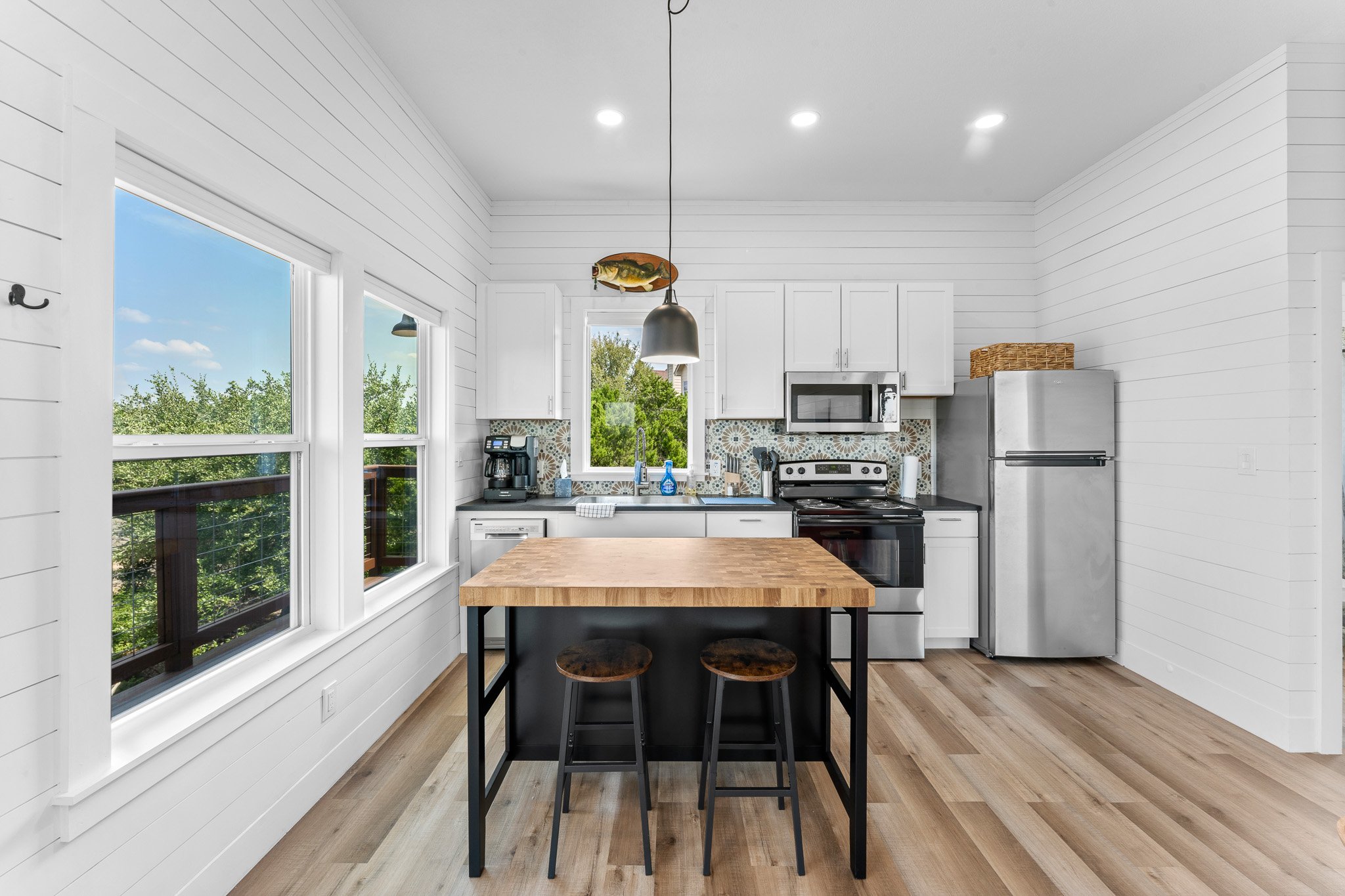 Modern kitchen with white shiplap walls, three large windows showing green trees outside, stainless steel refrigerator, microwave, coffee maker, and colorful backsplash. Dark wood table with matching stools in the center, floating fish decoration on 