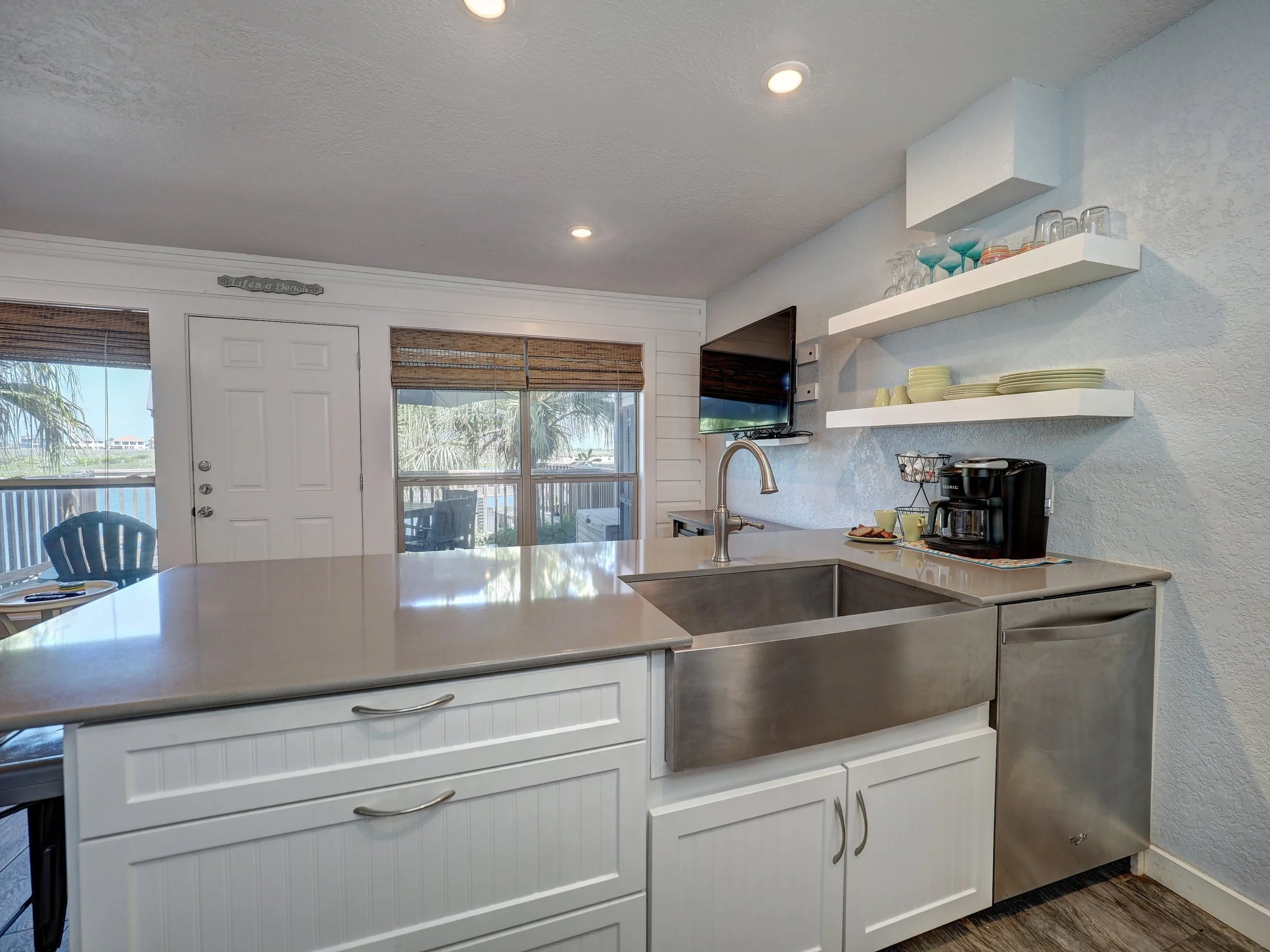 Kitchen with white cabinetry, a gray countertop, mounted shelves with glasses and dishes, stainless steel appliances, and a view of a patio with outdoor chairs and greenery outside large windows.