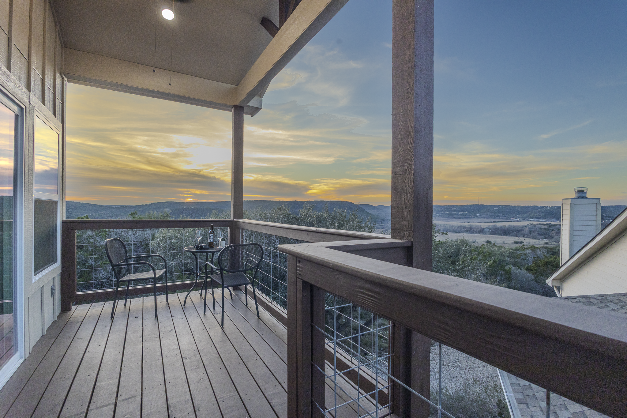 A wooden balcony with a metal railing and four black chairs, overlooking a scenic landscape with hills and a sunset sky.