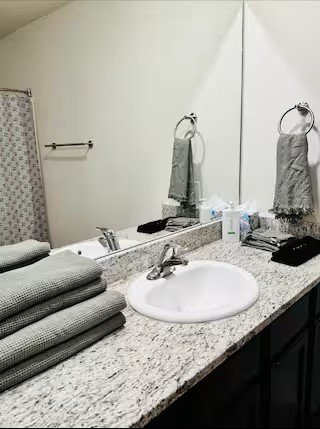 Bathroom sink with folded gray towels on granite countertop, mirror, and towels hanging on towel rings.