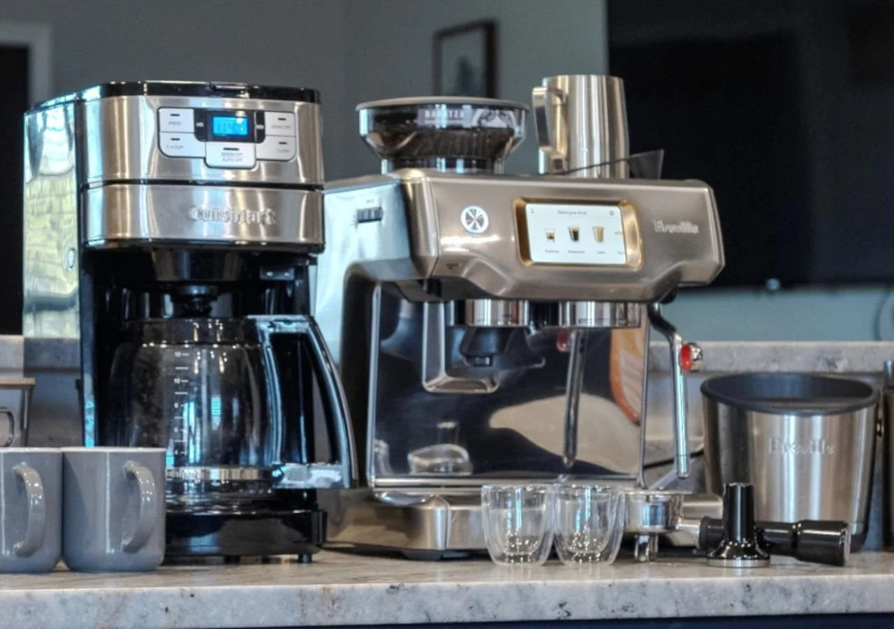 A stainless steel coffee machine with a built-in grinder, a digital display, and a drip coffee pot. There are two gray mugs, three small clear glasses, and a milk frother on the counter.
