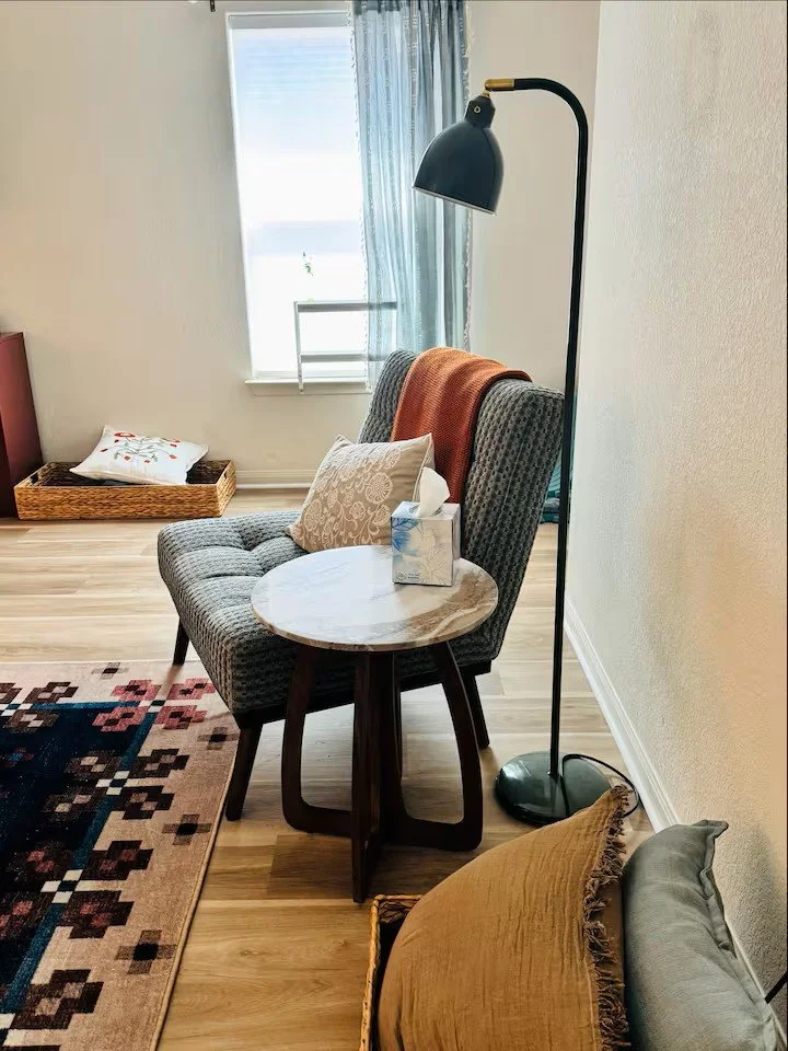 A cozy living room corner with a checkered armchair, a pillow, a small side table with a tissue box, a tall floor lamp, a window with curtains, and a dog bed with a pillow on the floor.