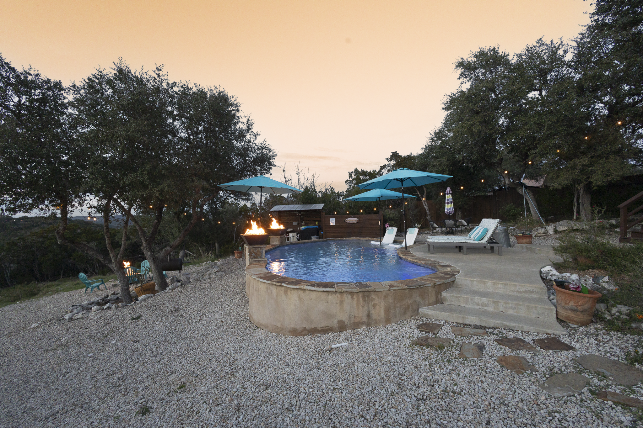 A backyard scene at sunset featuring a small pool with a stone border, surrounded by lounge chairs and patio furniture under blue umbrellas. A fire pit is lit, and string lights are hanging among the trees, creating a cozy outdoor atmosphere.