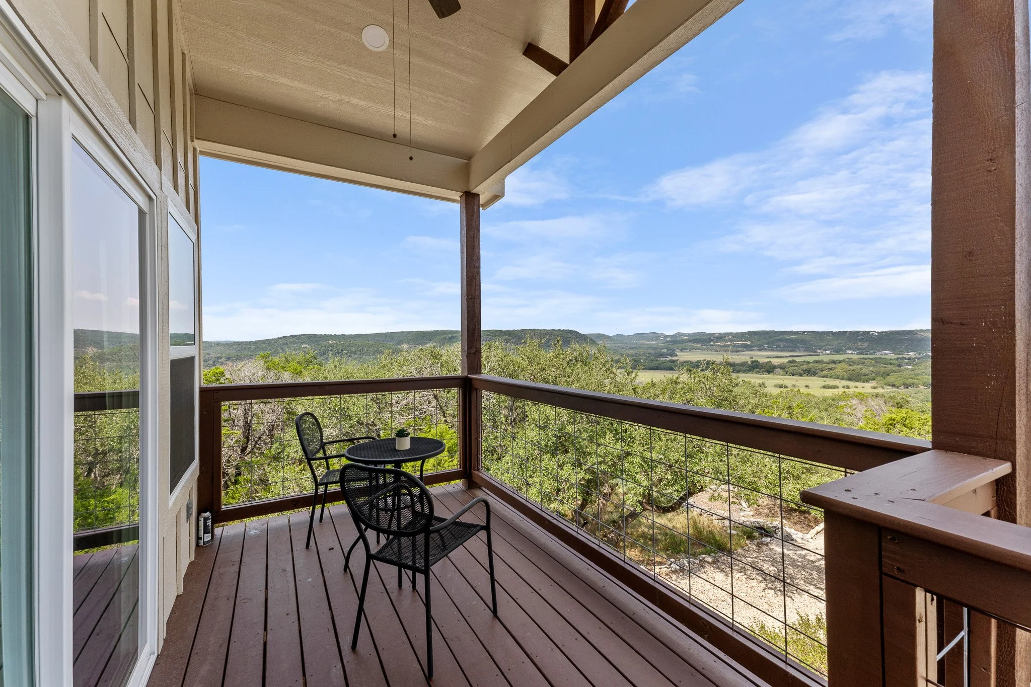 A balcony with black chairs and a small table overlooking a view of green trees and rolling hills under a partly cloudy sky.