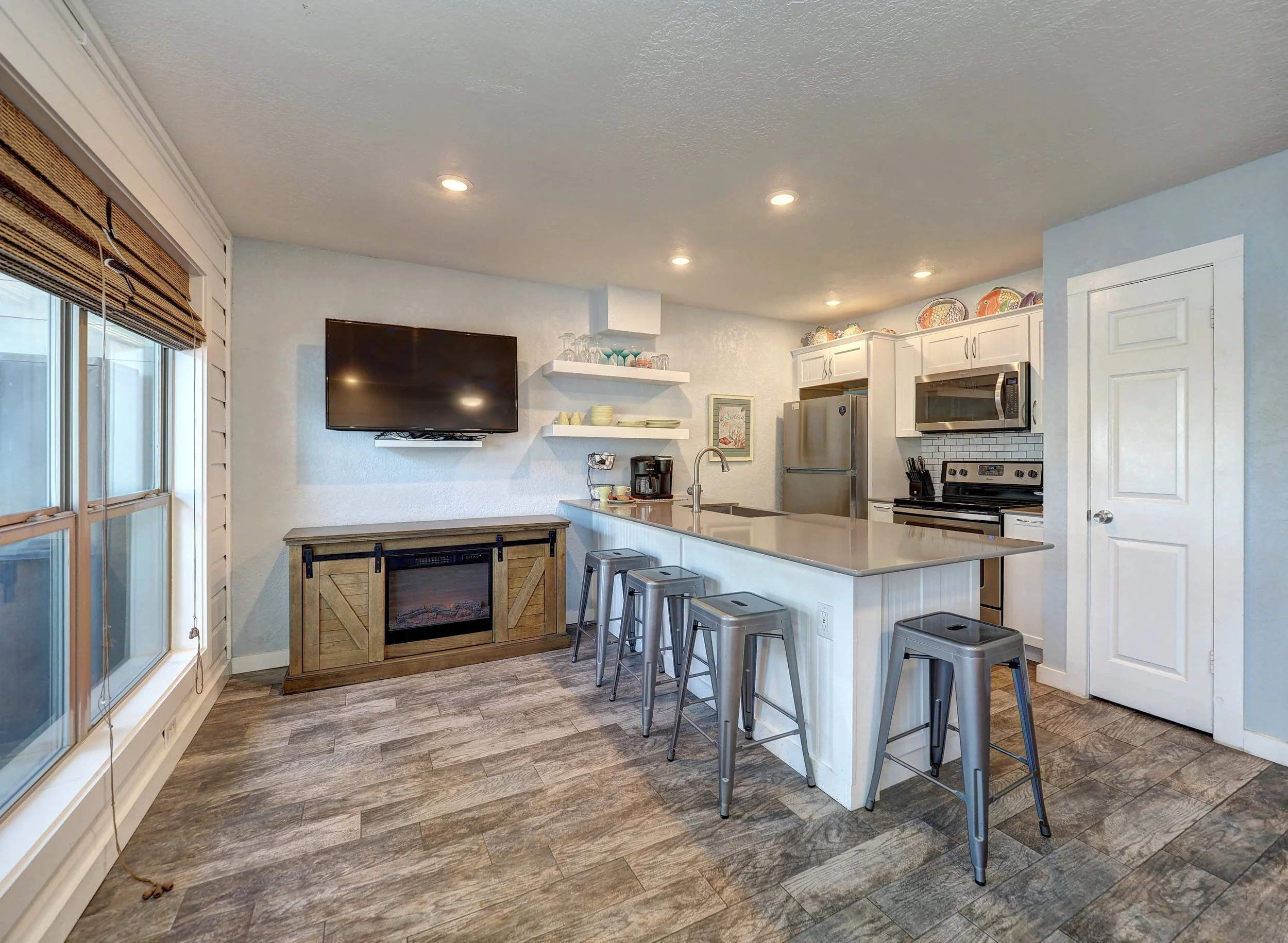 A kitchen with white cabinets, stainless steel appliances, a large island with a sink and four metal bar stools, a wall-mounted TV, floating shelves with dishes and glassware, a window with blinds, and brown wood-look flooring.