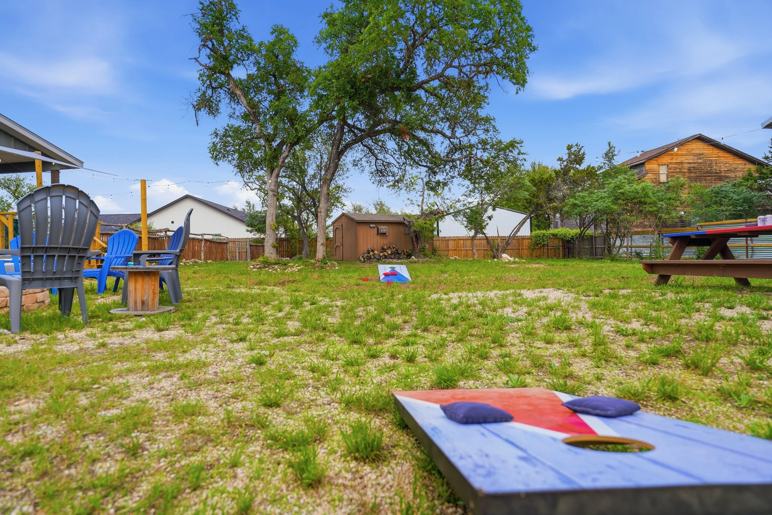A backyard with green grass, large trees, and various outdoor furniture including Adirondack chairs and a picnic table, with a shed and neighboring houses in the background.