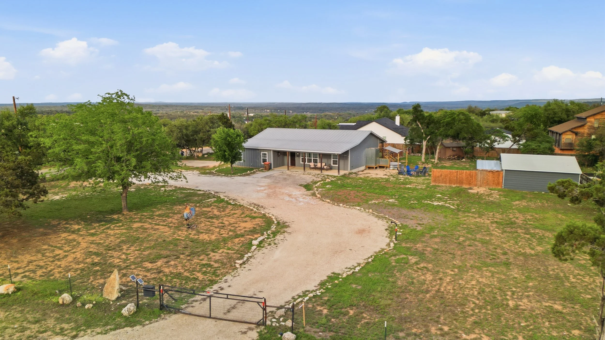 A rural residential scene showing a gravel driveway leading to a blue house with a covered porch, surrounded by green trees, with other houses and open landscape in the background under a partly cloudy sky.
