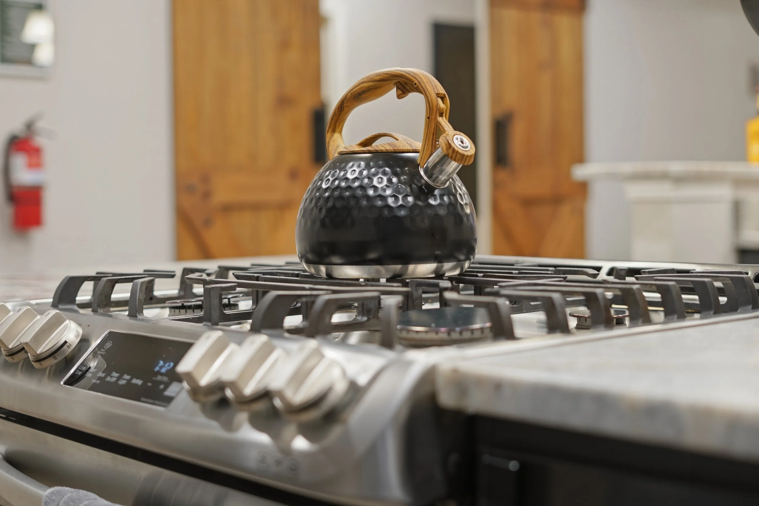 A black kettle with a wooden handle and a small wooden knob on the lid, placed on a stainless steel stove with control knobs visible in the foreground. The kitchen background includes wooden cabinets, a fire extinguisher on the wall, and a light-colo