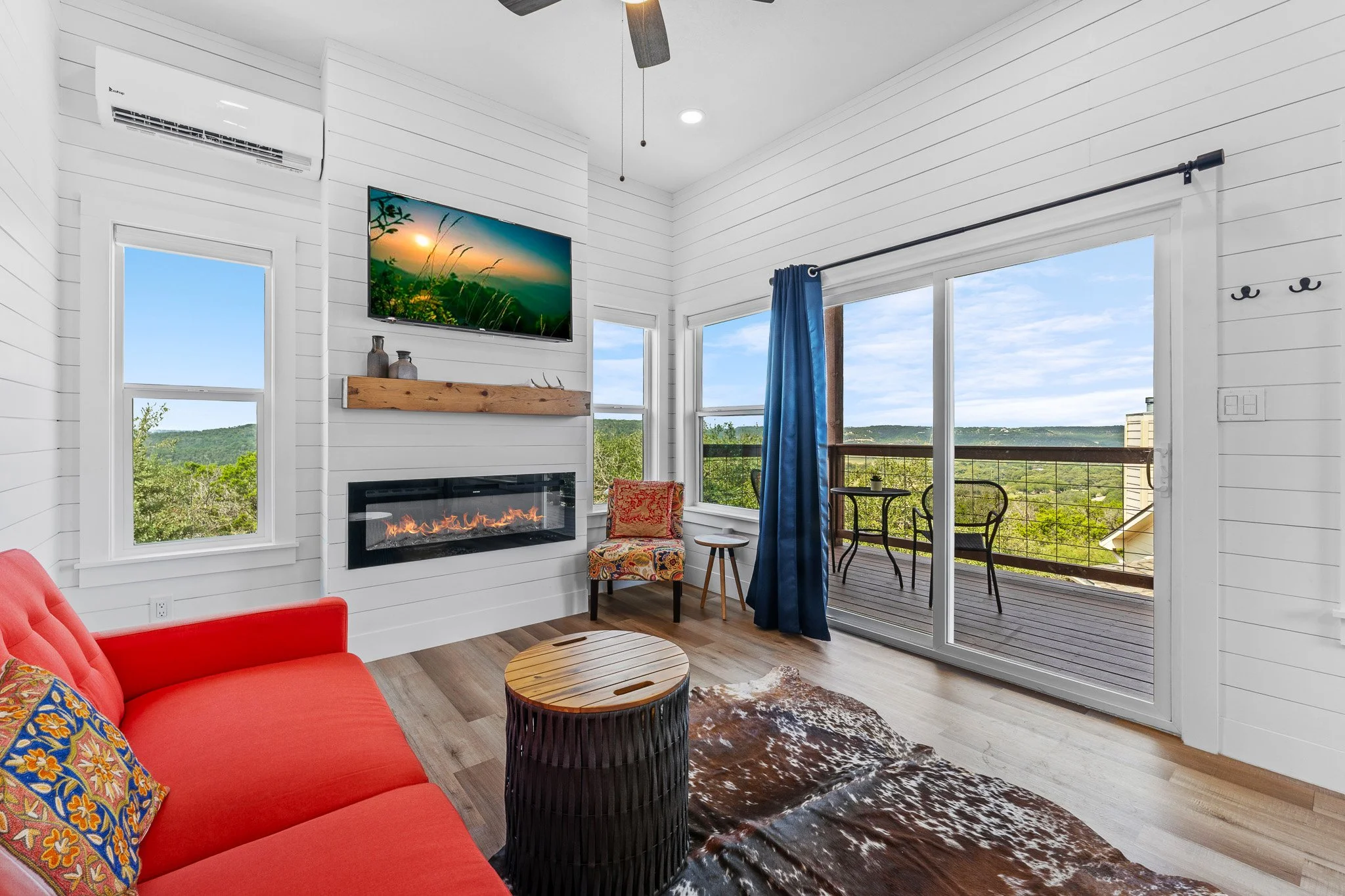 Living room with white shiplap walls, a red sofa with colorful throw pillows, a wooden coffee table, a cowhide area rug, a fireplace, a wall-mounted TV, and large windows with a view of a balcony and green landscape outside.