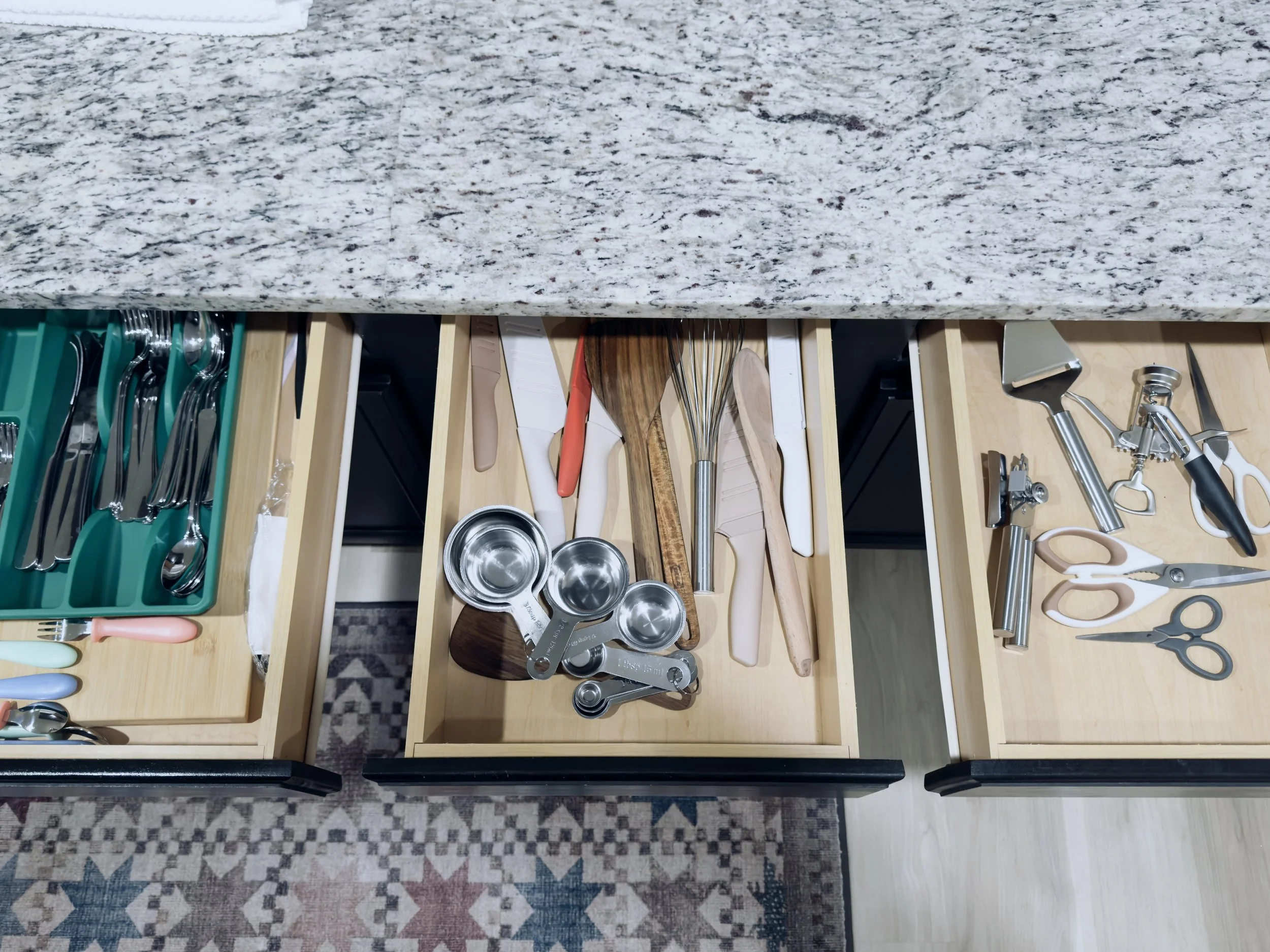 Open kitchen drawers containing various kitchen utensils and tools, including measuring cups, wooden spoons, silicone spatulas, and metal whisks, with the drawers positioned beneath a granite countertop.