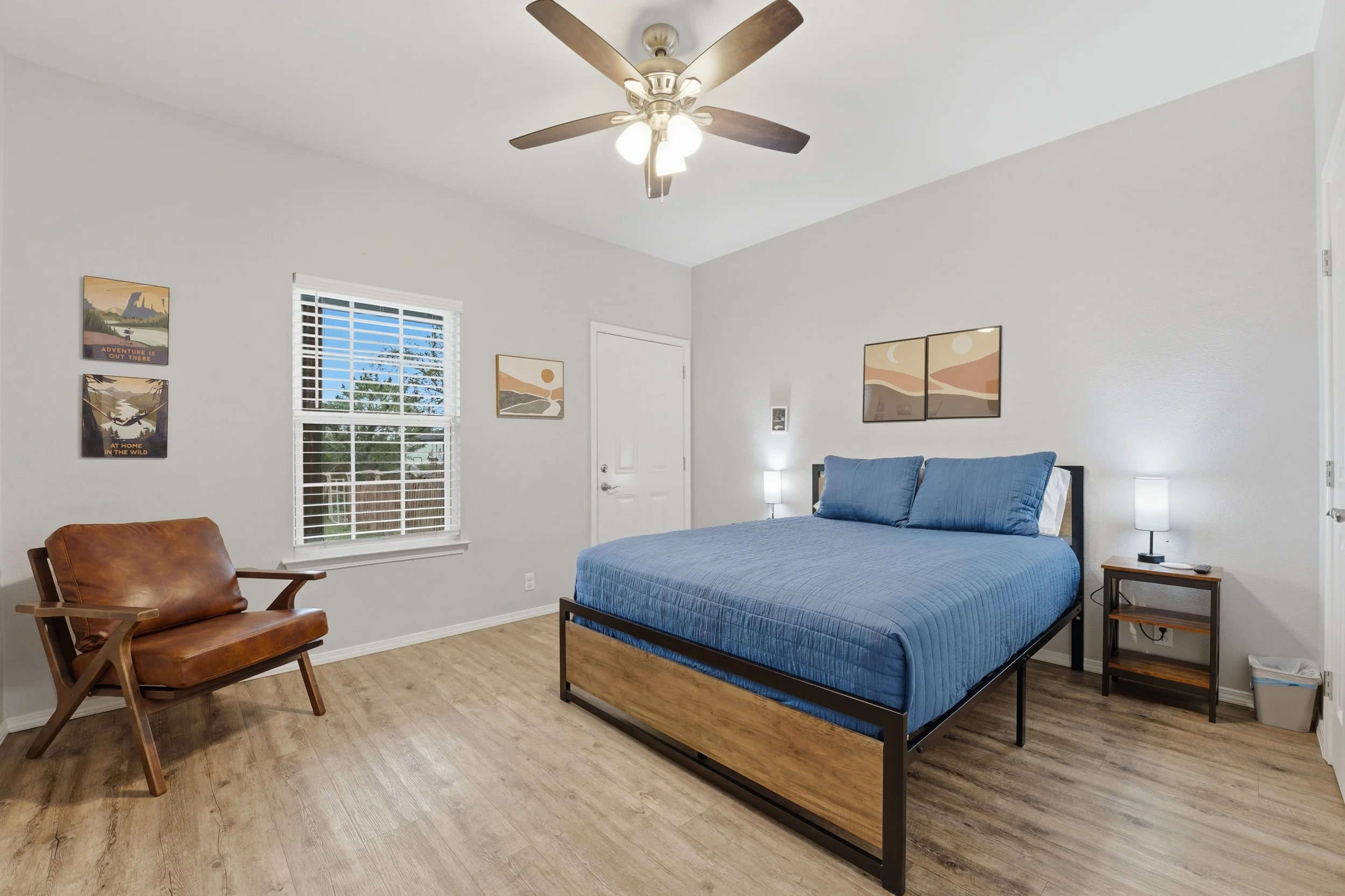 A bedroom with a bed featuring a blue quilt, a wooden and metal frame, and two side tables with lamps. There is a leather armchair near a window with white blinds, and three framed landscape posters on the walls. The ceiling has a fan with wooden bla