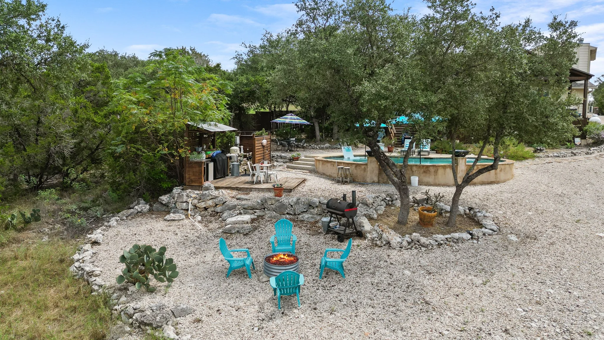 A backyard with a circular fire pit surrounded by blue Adirondack chairs, a gravel area, trees, a hot tub, and outdoor furniture with an umbrella.