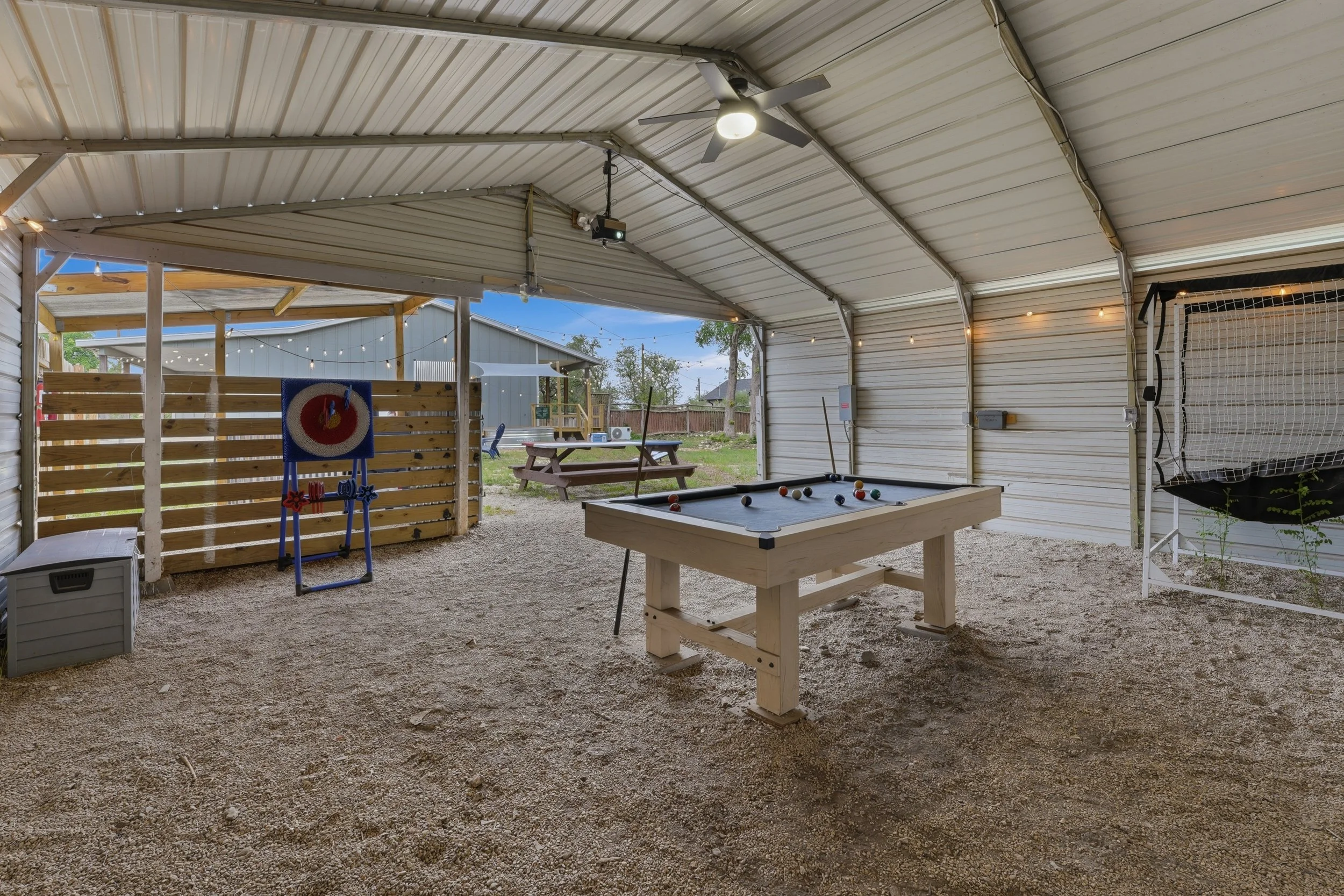 A covered outdoor space with a ceiling fan, string lights, a shuffleboard table, and a pool table. There is a wooden privacy wall, a picnic table, and a badminton net. The area opens to a backyard with a shed and trees.