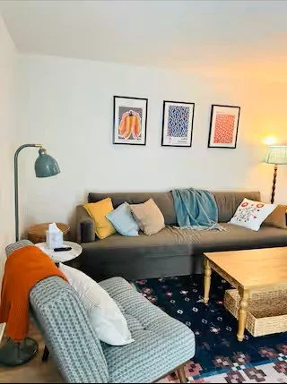 Living room with a gray sofa decorated with yellow, blue, and white pillows, blue throw blanket, and framed artwork on the white wall. There's a patterned armchair with white pillows, a wooden coffee table, a floor lamp, and a small side table with a