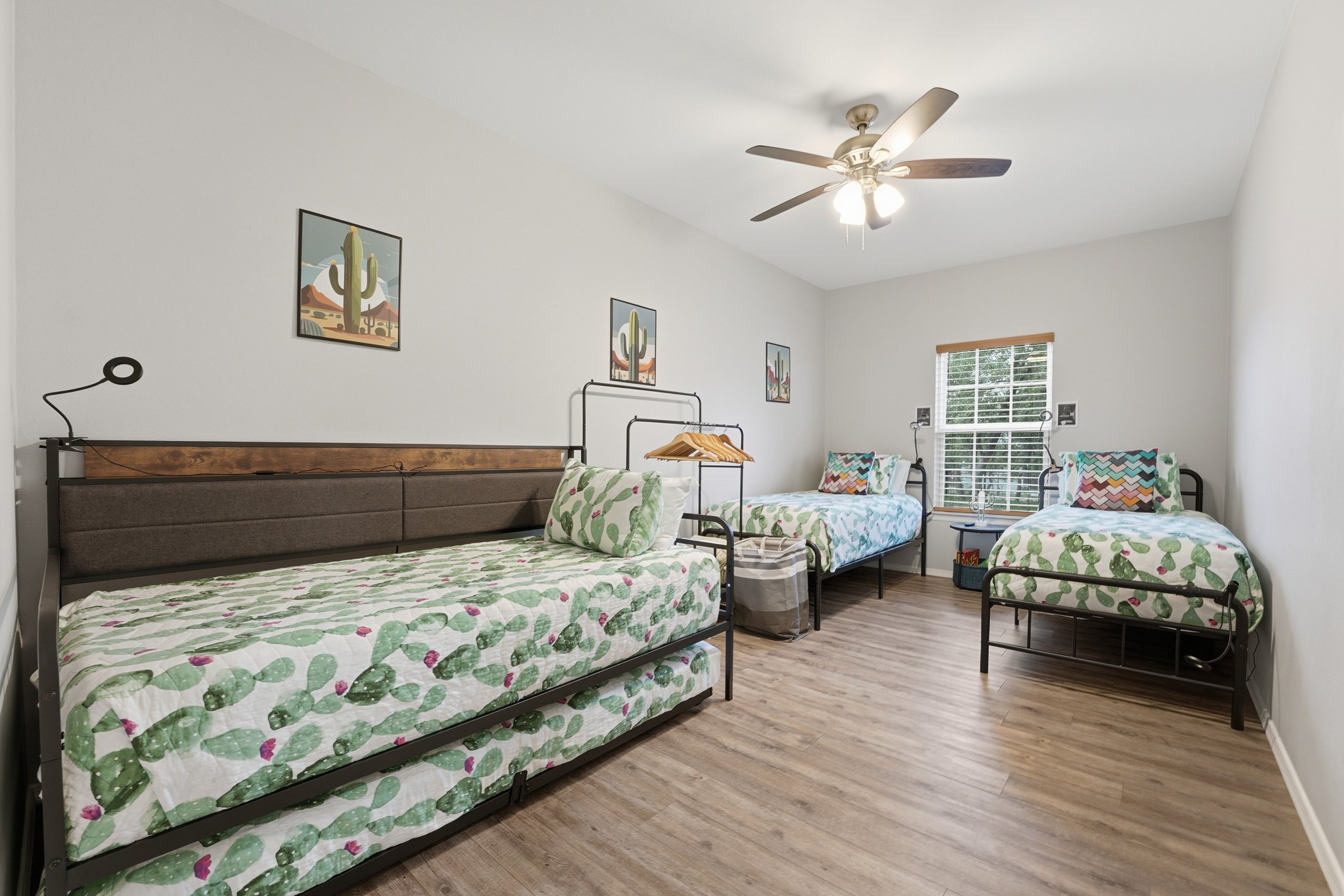 A brightly lit bedroom with three beds, each with floral bedding, wooden flooring, and a ceiling fan. The walls are decorated with cactus artwork, and there is a window with blinds.