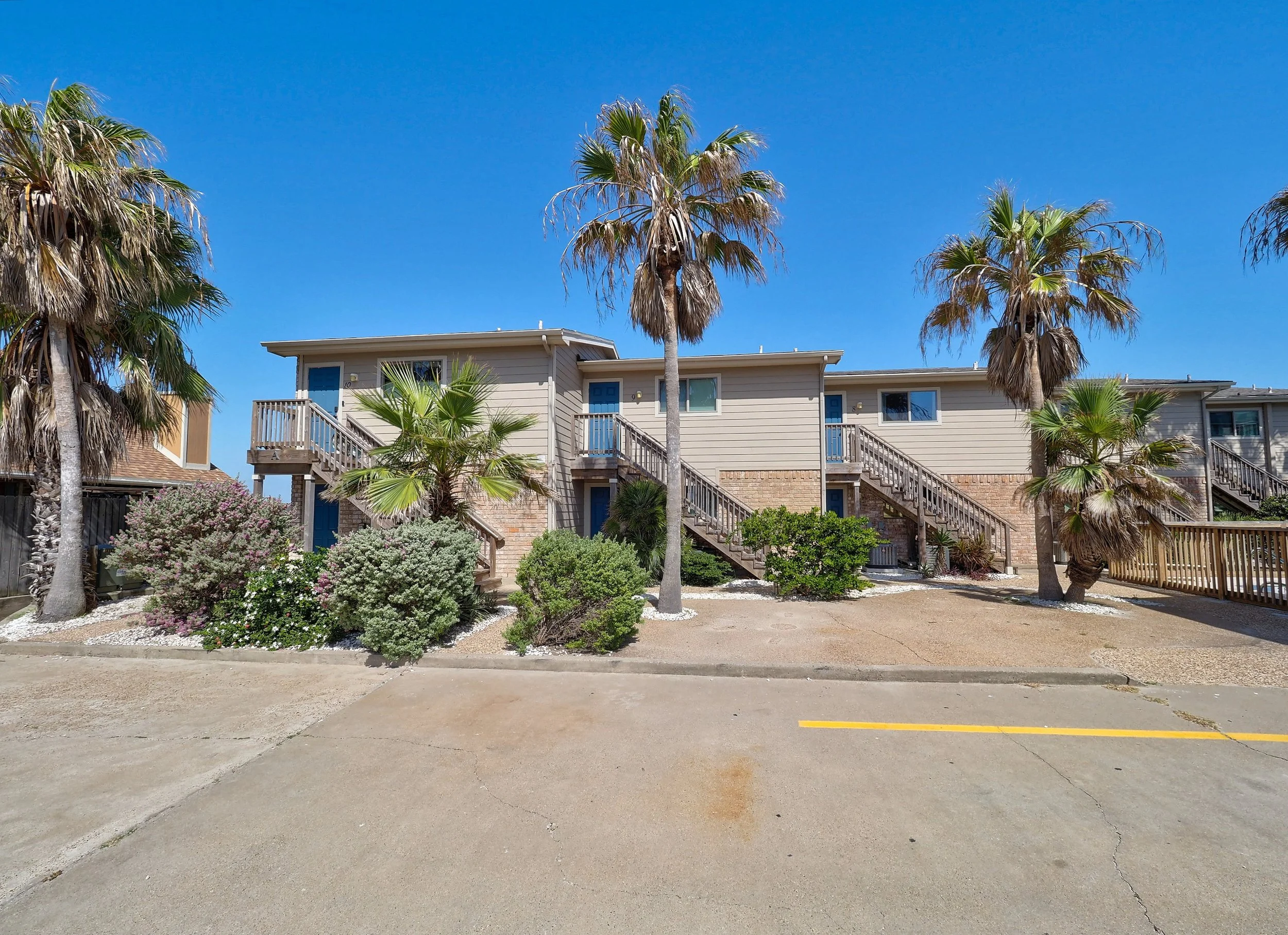 Apartment building with palm trees and shrubs in front, clear blue sky, and a parking lot with yellow line.
