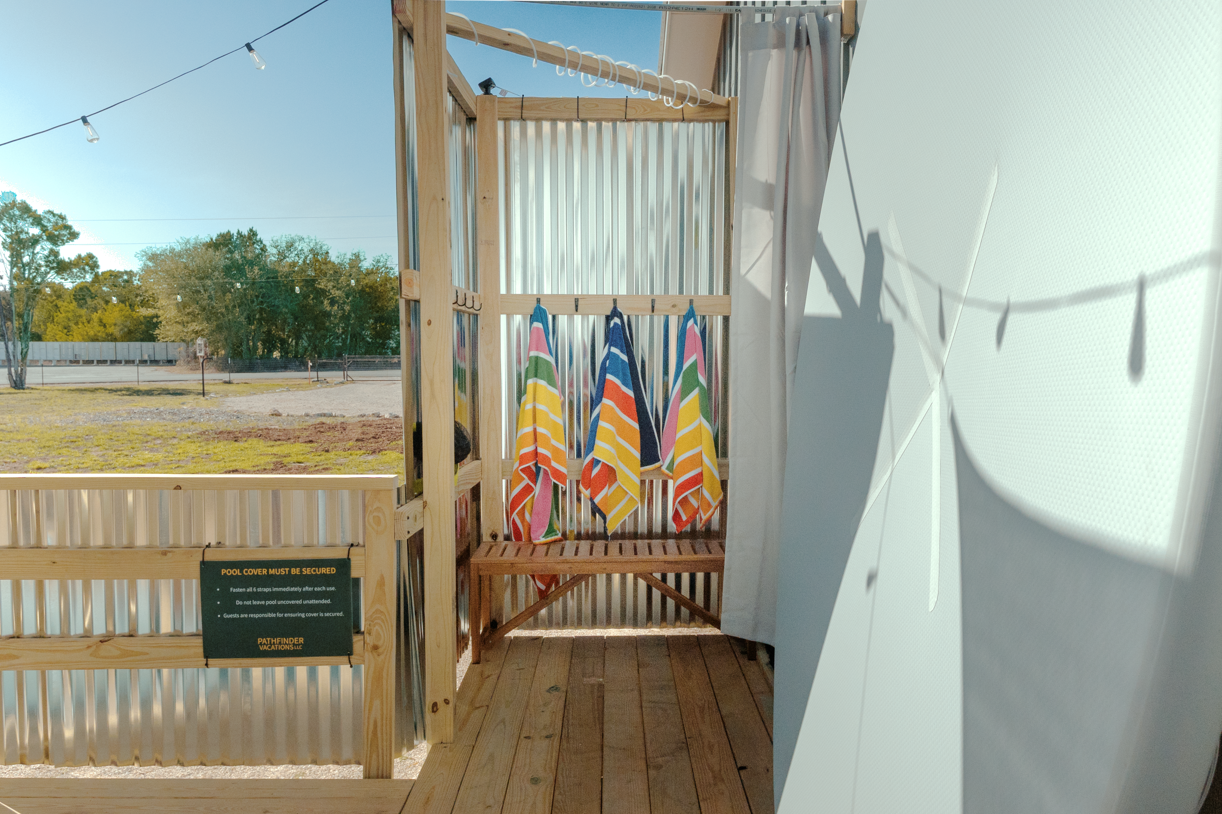 A small outdoor shower area with a wooden platform and a metal privacy screen. The shed is decorated with six colorful towels hanging on a horizontal rod, under a wooden overhead bar. Sunlight casts shadows of the towels and other objects onto the white wall.