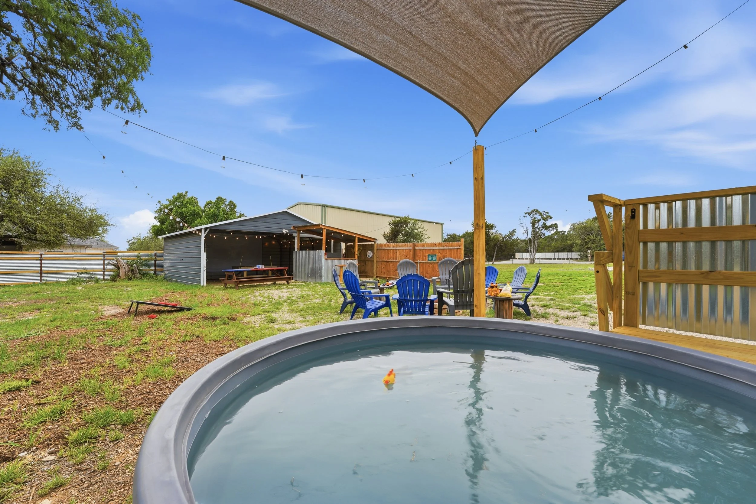 Backyard scene with a round inflatable pool in the foreground, a patio table with chairs, string lights, a wooden fence, a small shed, trees, and a clear blue sky with a shade sail overhead.