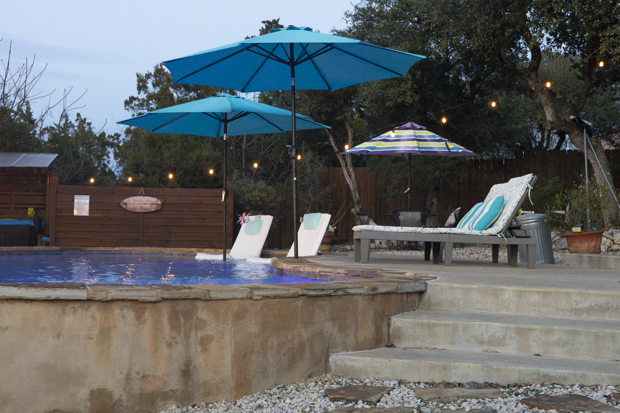 Backyard pool area with three blue umbrellas, a lounge chair with pillows, a sofa, and string lights in the trees at dusk.